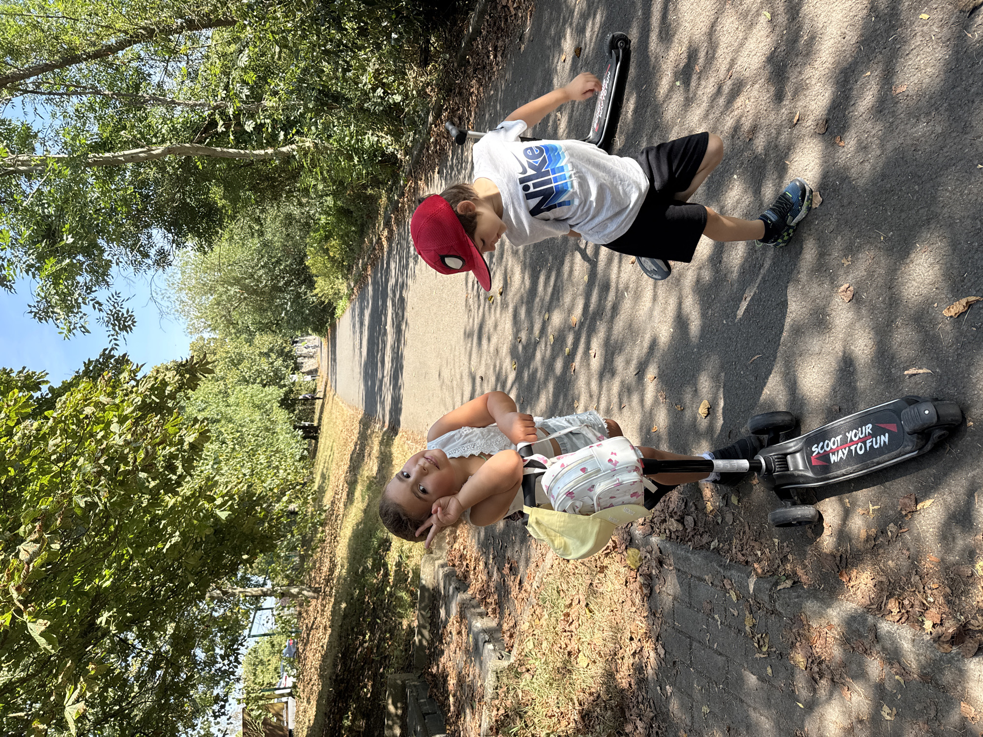 In an outdoor scene on a paved path bordered by trees and grass, two young children are present. On the left, a girl stands with a three-wheeled scooter, making a peace sign with her right hand. She wears a white lace top, light-colored shorts, and has a yellow cap and a patterned backpack on her scooter. On the right, a boy is on his own scooter, looking down. He is wearing a red baseball cap with a spider emblem, a white Nike t-shirt, and black shorts. The path appears to be in a park or a tree-lined street, with sunlight dappling through the leaves. The environment suggests a warm, sunny day. The image does not contain any information or visual cues that can be used to determine its location within the city of Clonmel, Ireland.