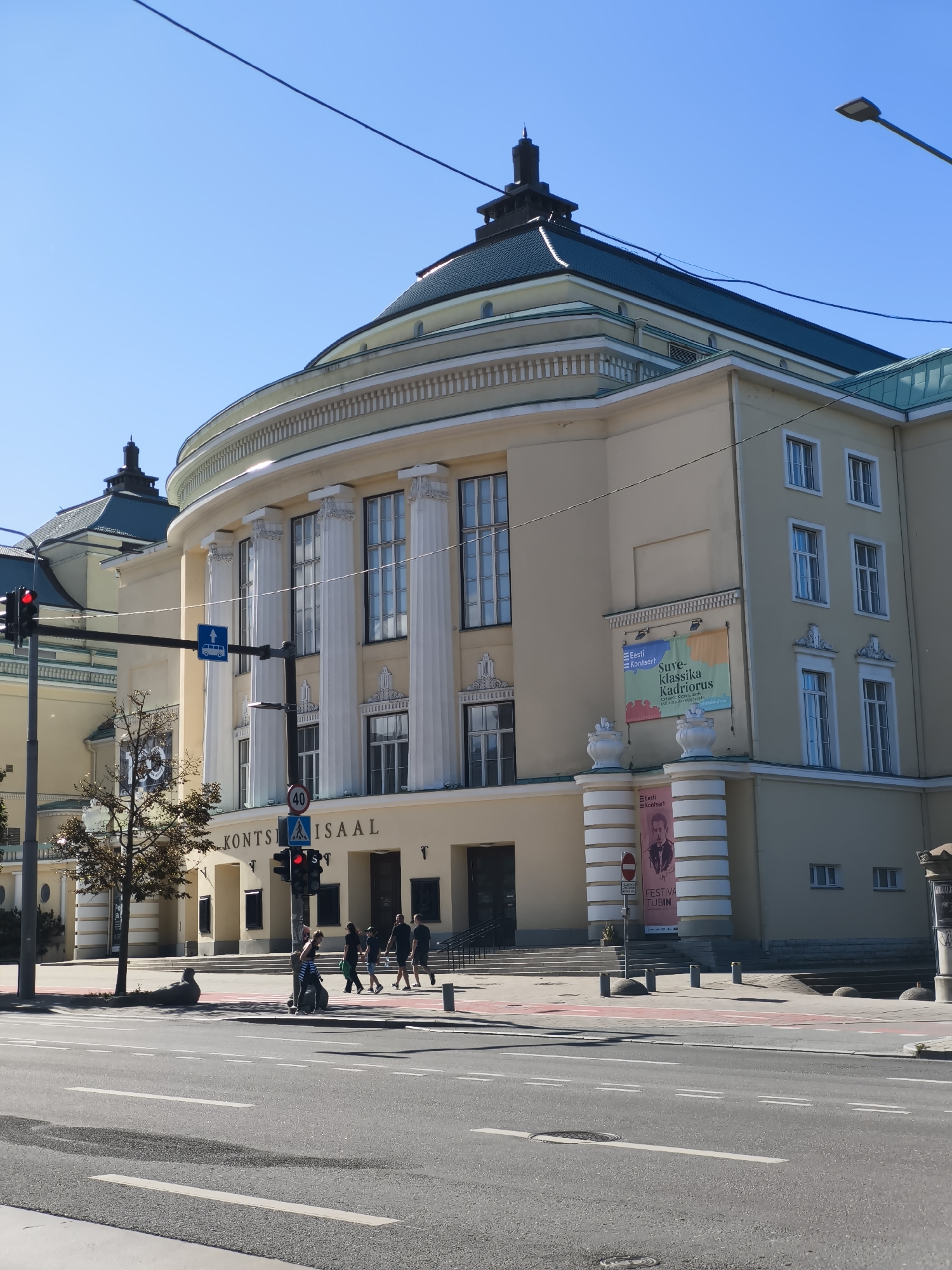 The image shows a street scene in Tallinn, Estonia, with the Estonia Concert Hall prominently featured. The building, with its light yellow facade and classical architectural elements like columns and arched windows, dominates the upper portion of the frame. A dark teal-colored roof caps the structure, and a distinctive spire is visible on one of the building's sections.
In the foreground, a street with multiple lanes of asphalt is depicted, marked with white lane lines and a red-painted bike lane. Traffic lights are visible, one showing red. Several people are walking on the sidewalk in front of the concert hall, some carrying bags. A few trees are planted along the sidewalk, with one particularly prominent tree in the left half of the image. Advertisements are displayed on the facade of the Estonia Concert Hall, including one with a portrait of a man and text that appears to be in Estonian. A speed limit sign showing "40" is also visible. The sky is clear and blue.