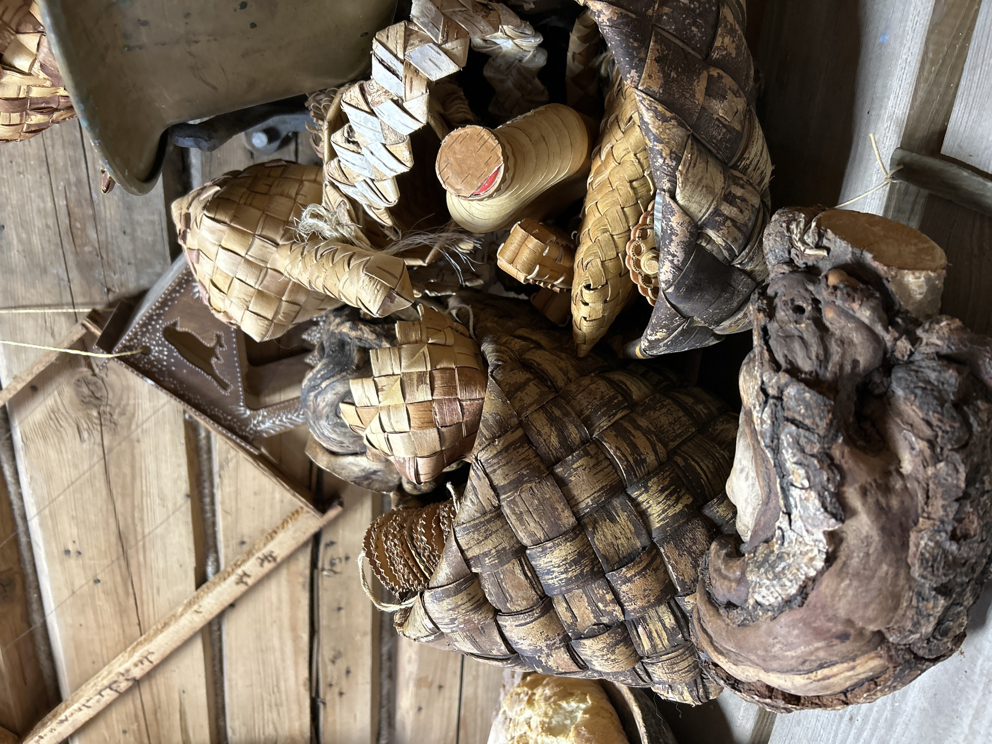 The image depicts a collection of woven birch bark objects and wood fragments indoors. The objects vary in size and shape, including baskets, containers, and decorative items. Some are made entirely of woven bark, while others incorporate wood components. The bark exhibits different tones of brown and cream. The objects are arranged on a wooden surface, possibly a table or shelf. A metallic object is visible in the upper left corner. The location is Moscow, Russia.