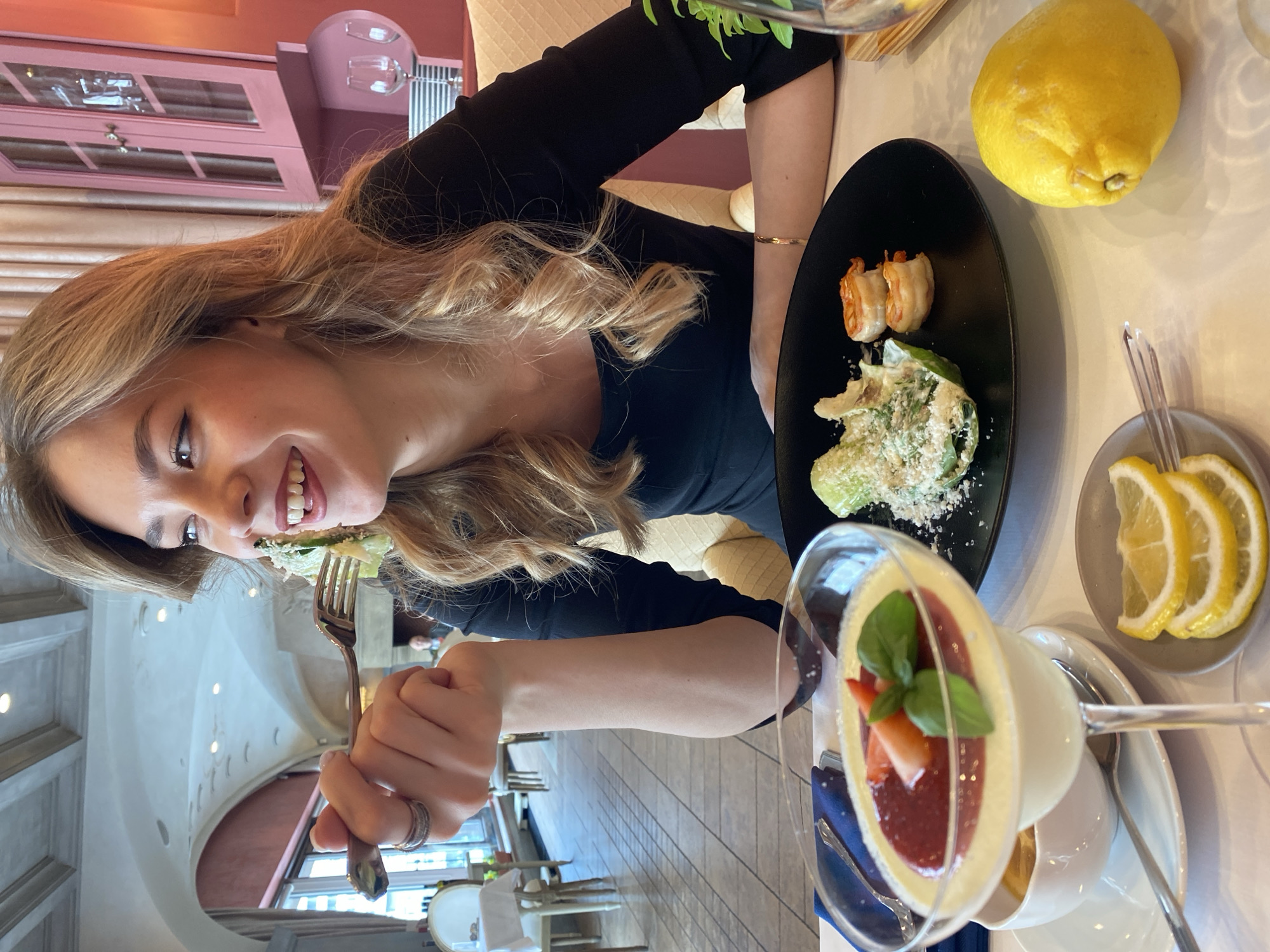 The image, rotated 90 degrees counter-clockwise, depicts a young woman smiling joyfully while dining in a restaurant in Yekaterinburg, Russia. She has long, wavy blonde hair and wears a black long-sleeved top. Her left hand holds a fork with a piece of salad, poised near her mouth. Rings are visible on her fingers, and a thin bracelet adorns her right wrist.
On the table in front of her, a black oval plate holds two pieces of what appear to be wrapped shrimp and a green salad generously topped with grated cheese. Beside the plate, a large, whole yellow lemon is placed directly on the table, along with a small grey dish containing three fresh lemon slices. A dessert is prominently featured in a stemmed glass, showcasing a creamy layer topped with a vibrant red fruit sauce, garnished with fresh strawberry pieces and basil leaves. A small white pitcher and a spoon accompany the dessert.
The background reveals the interior of an elegant restaurant or cafe. Notable architectural features include a light-colored, arched ceiling with decorative lighting fixtures. Some walls and cabinetry have a reddish-pink hue. Ornate white chairs and tables with white tablecloths are visible in the background, suggesting a refined dining atmosphere. The floor is tiled. No visible text is present in the image.