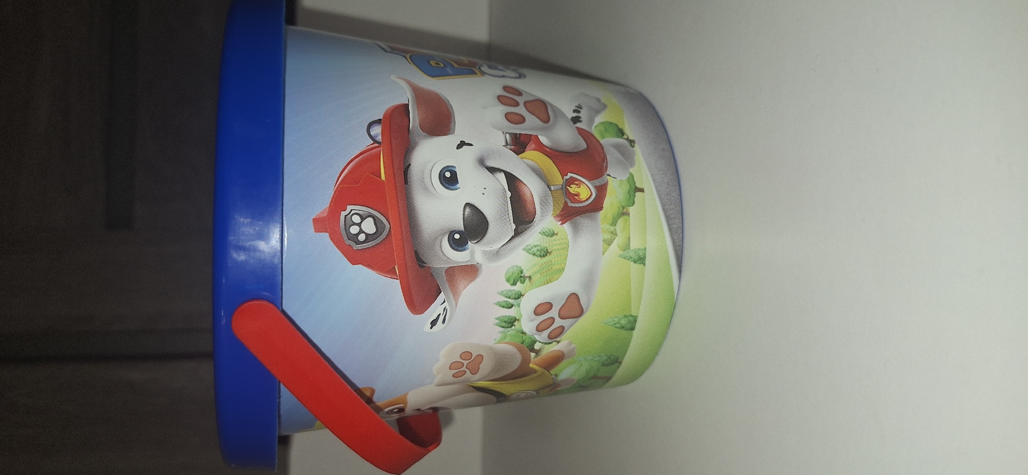 The image shows a close-up of a blue-topped plastic bucket decorated with characters from the children's show Paw Patrol. The bucket is predominantly light blue and features a scene with two cartoon dogs wearing firefighter gear. One dog, Marshall, is prominently displayed in the foreground, smiling and waving. The other dog, likely Rubble, is visible on the left. The background depicts a stylized green landscape with trees under a bright blue sky. The bucket has a red handle attached to the blue rim. The object is placed on a white surface, with a dark, out-of-focus background suggesting an indoor setting. There is no information in the image that can identify its location in Bila Tserkva, Ukraine.