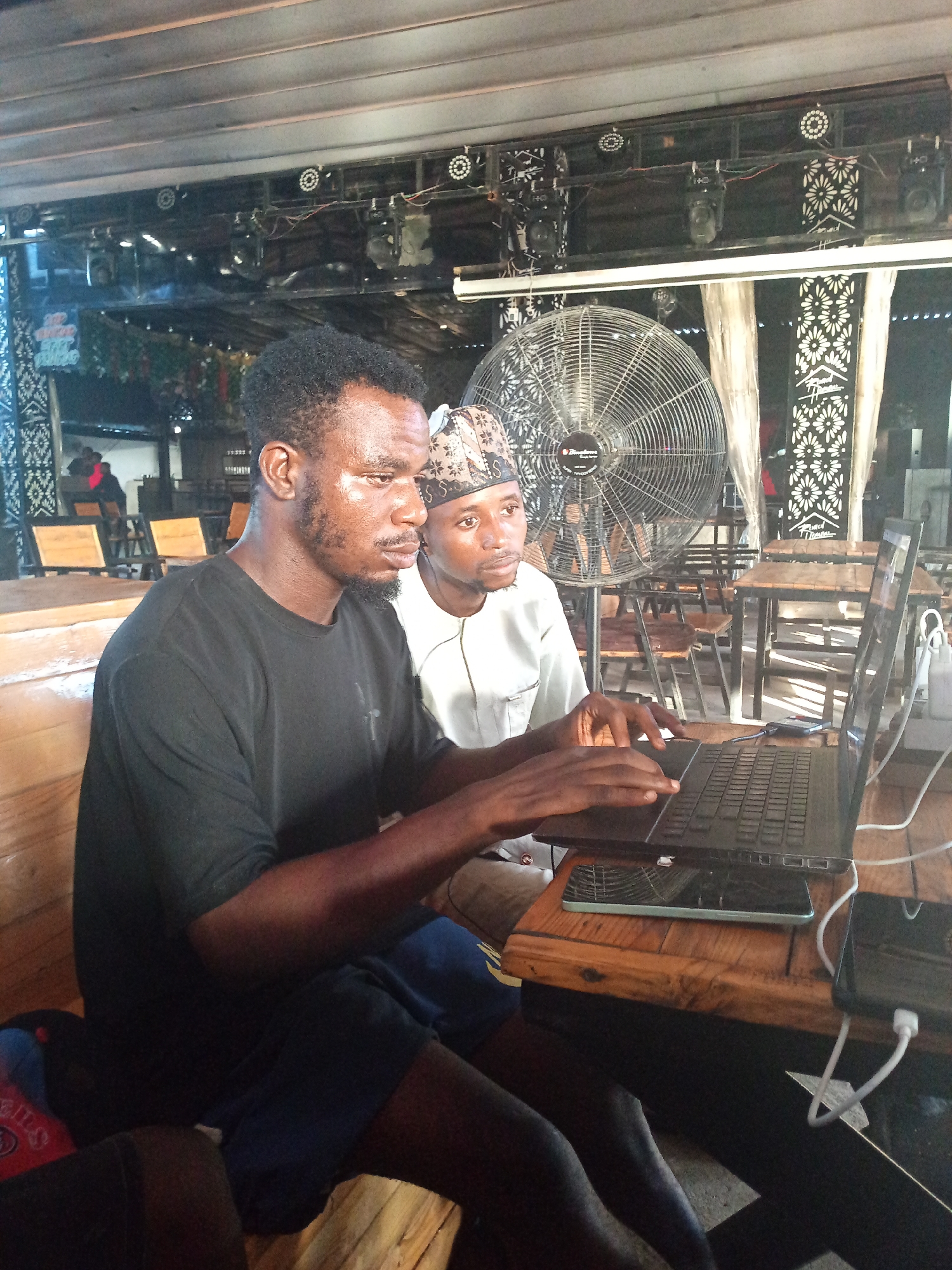 This image depicts two males engaged with a laptop at a wooden table in an indoor or semi-open-air venue in Howayi, Nigeria. The male on the left, with dark skin, a beard, and short dark hair, wears a black t-shirt and dark shorts, and is actively typing on a black laptop. Beside him, a second male, also with dark skin, wears a light-colored traditional top and a patterned cap, observing the laptop screen. On the dark wooden table, an open black laptop is positioned, with at least two smartphones nearby; one lies partially beneath the laptop with a visible green/blue casing, and another is to the right, connected to white charging cables and an adapter. In the background, a large standing "Brandson" fan with a metal cage is visible. Above, a ceiling structure features a black frame with multiple circular and spotlight-style lights, interspersed with decorative panels displaying a white floral pattern on a dark background. One such panel contains the text "Grand House". Additional wooden tables and chairs, along with indistinct figures of other individuals, are present in the deeper background.