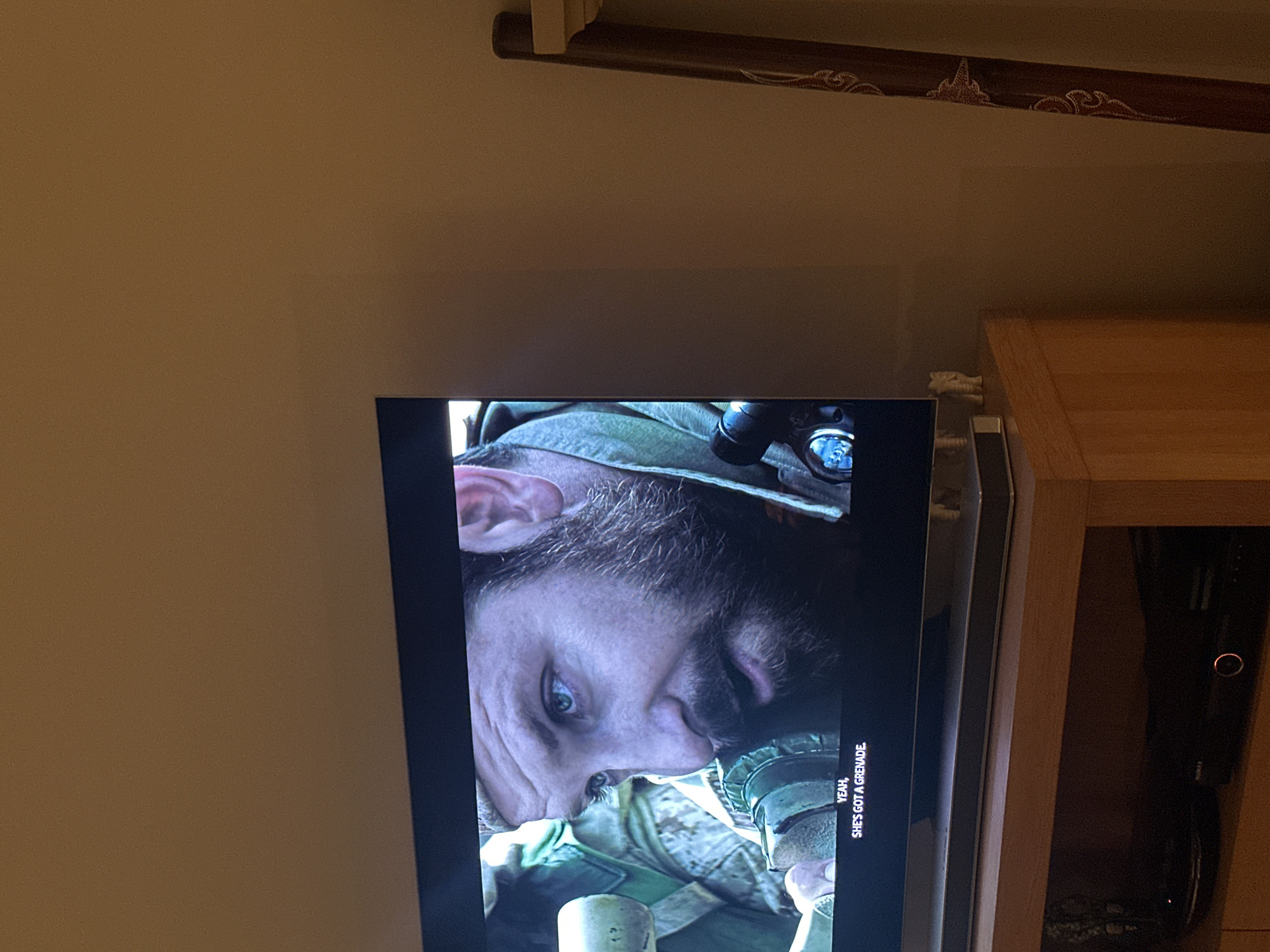 The image shows a close-up of a television screen displaying a scene from what appears to be a war-themed video game or film. A male character, wearing military gear and night vision goggles, is prominently featured. Text on the screen reads, "Yeah, she's got a grenade."
The television is positioned in front of a wooden cabinet or shelving unit. The background includes a portion of a wall, and a decorative wooden trim detail is visible near the top of the frame. There is no location information within this image.