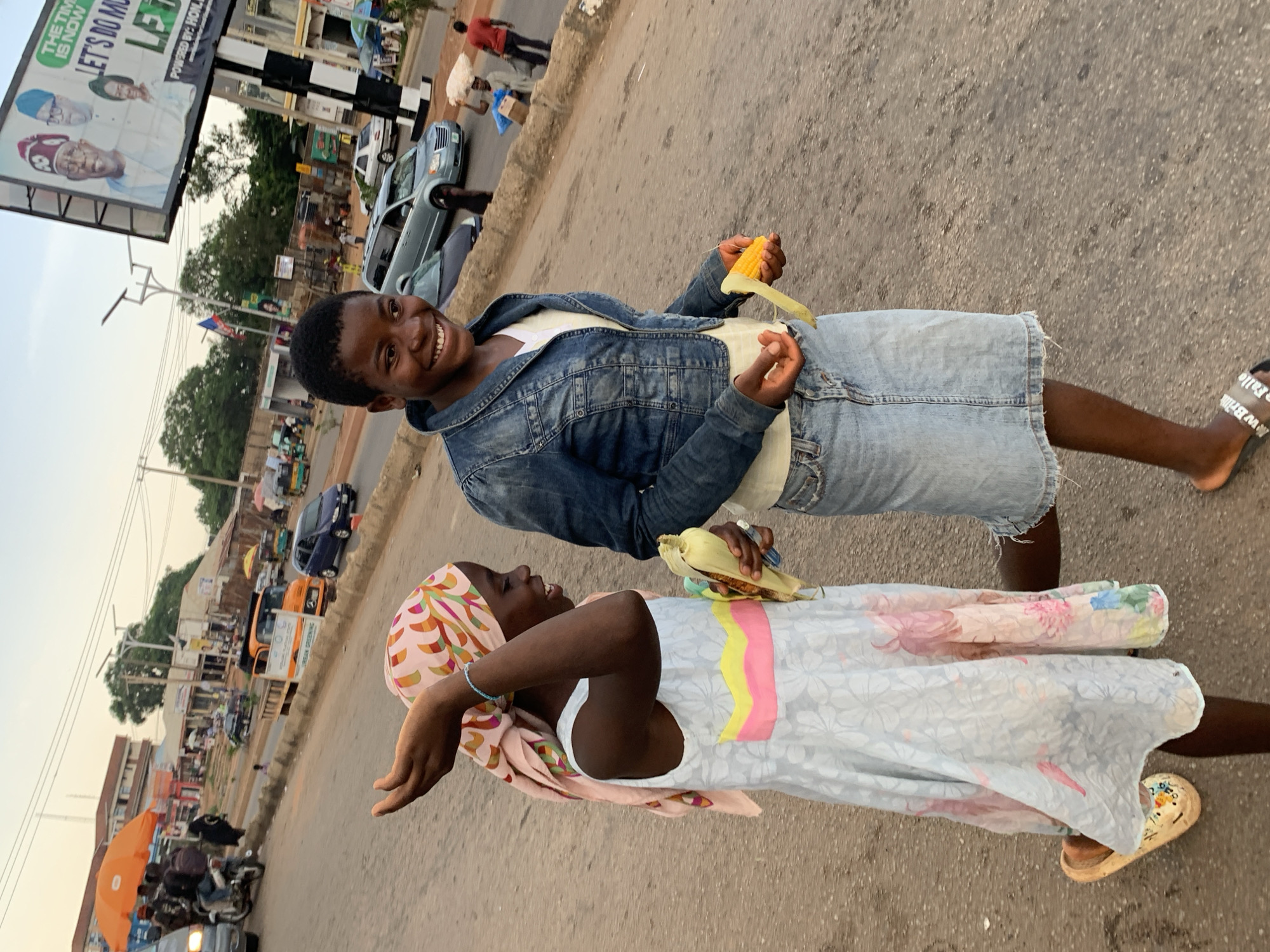 The image depicts two young girls walking on a street in Jos, Nigeria. The girl on the left wears a light blue dress with floral patterns and a pink headscarf. She is holding a piece of corn in her right hand and appears to be reaching out with her left. The girl on the right is wearing a denim jacket and a denim skirt, and is also holding a piece of corn. Both girls are smiling and appear to be interacting with each other. The background shows a street with traffic, including cars, motorcycles, and pedestrians. A large billboard with political figures is visible on the right side of the image, suggesting a public area in the city. The overall atmosphere suggests a casual, everyday scene in Jos.