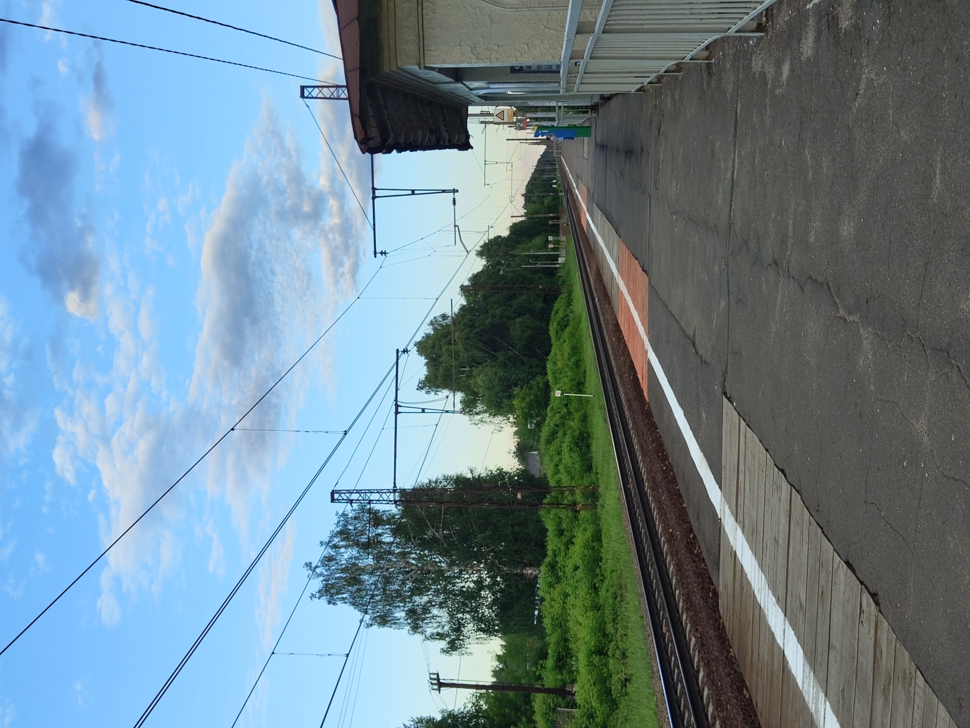 This is a ground-level, eye-level, static, medium shot of a train platform and tracks at a train station in the city of Tsibino, Russia. The train tracks run horizontally across the image from left to right, with ballast underneath. A concrete platform with a white line and an orange painted section runs alongside the tracks. A wooden ramp crosses the platform perpendicular to the tracks. To the right of the platform, a light-colored building with a brown roof and white railings is visible. Overhead, numerous power lines for the electric train are strung between tall poles and crisscross the sky. The sky is partly cloudy with a mixture of blue and white clouds. Lush green trees and bushes line the area beyond the tracks. The overall scene depicts a rural or suburban train station.