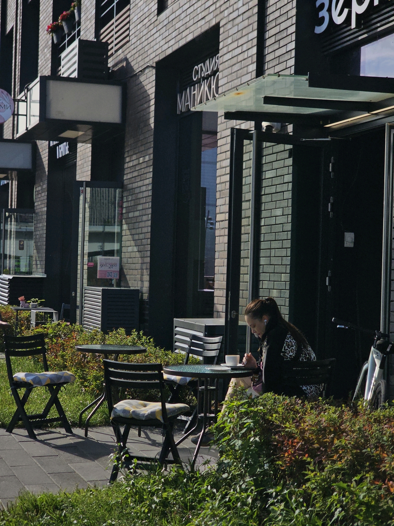 The image depicts an outdoor seating area of a cafe in Moscow, Russia. A woman with dark hair pulled back in a ponytail sits at a small round table, wearing earphones and a leopard-print jacket. She is looking down at her hands, which are near a white mug and saucer on the table. In the background, a brick building houses a cafe with a sign that partially reads "СТУДИЯ МАНИКЮР" (Studio Manicure). A bicycle with a white frame is parked to the right of the cafe entrance. Several empty chairs with checkered cushions are arranged around other tables in the seating area, which is surrounded by green foliage and located on a paved walkway. The sun casts shadows on the ground, indicating it is daytime.