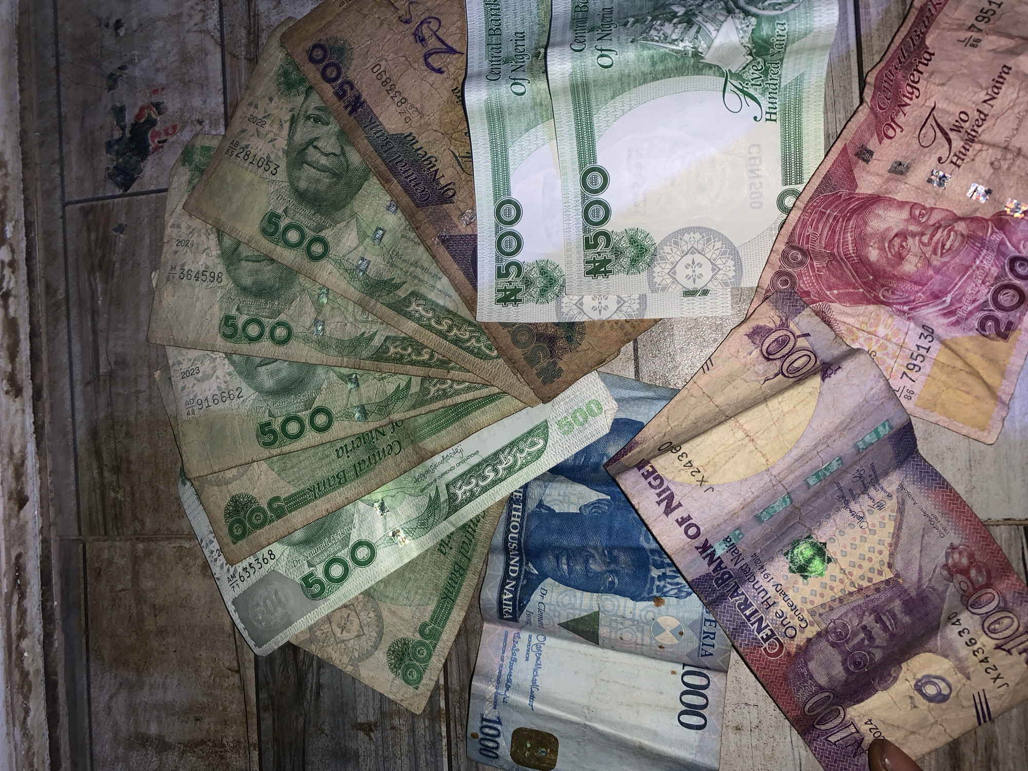 The image displays a collection of Nigerian Naira banknotes of various denominations spread out on a surface with a light woodgrain pattern. The majority of the banknotes are ₦500, featuring the portrait of Nnamdi Azikiwe. There are also several ₦1000 banknotes displaying the portraits of Dr. Clement Isong and Alhaji Aliyu Mai-Bornu, along with ₦200 banknotes bearing the image of Alhaji Sir Ahmadu Bello. Additionally, one ₦100 banknote depicting Chief Obafemi Awolowo is visible. The arrangement of the banknotes is somewhat disorganized, with some fanned out and others overlapping. The lighting in the image appears dim and uneven, causing varying shades across the scene. The image does not depict Jos, Nigeria, people, actions, or interactions.