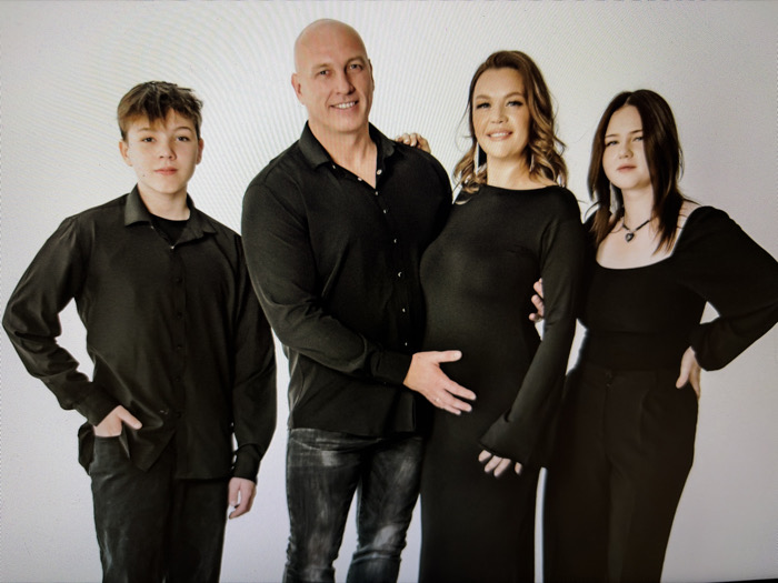 A family of four poses against a neutral backdrop. A bald man in a black button-down shirt stands next to a woman in a long black dress, whose pregnant belly he touches. To their left is a young man in a black shirt, hand in pocket. To the right is a young woman in a black square-neck top and trousers, wearing a necklace. The family is dressed formally and uniformly in black attire.