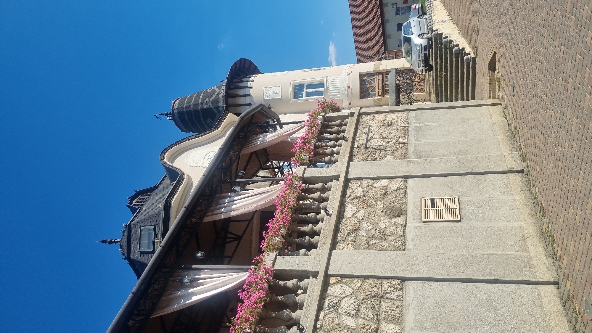 A street view in Berehove, Ukraine, features an Art Nouveau style building with ornate architectural details, including a conical tower and a curved roofline. A balcony runs along the side of the building, adorned with pink flowers in planters and draped light-colored curtains. The building's facade is a combination of stone and stucco. A silver sedan is parked on an elevated section of road next to a staircase leading up to the building's entrance. The sky is clear blue, and the ground is paved with brick.