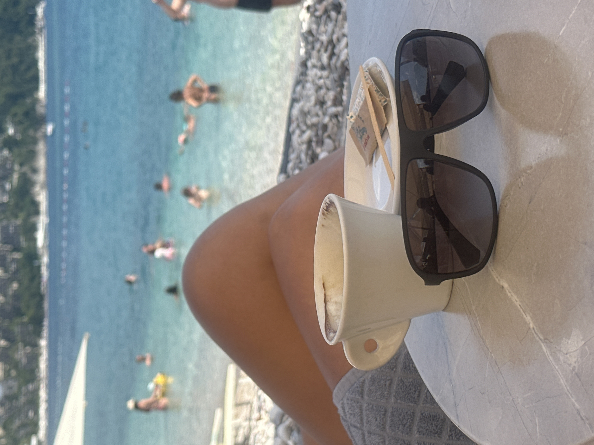 In Budva, Montenegro, a close-up shot focuses on a marble-topped table at a beachside cafe, where a white ceramic mug of coffee, a small white saucer with sugar packets and a wooden stirrer, and a pair of dark-rimmed sunglasses are arranged. In the background, the turquoise waters of the Adriatic Sea are visible, with several people swimming and enjoying the water. A rocky shoreline and a white beach umbrella complete the serene beach scene. The person whose legs are visible in the foreground is likely seated at the cafe, enjoying their coffee and the view.