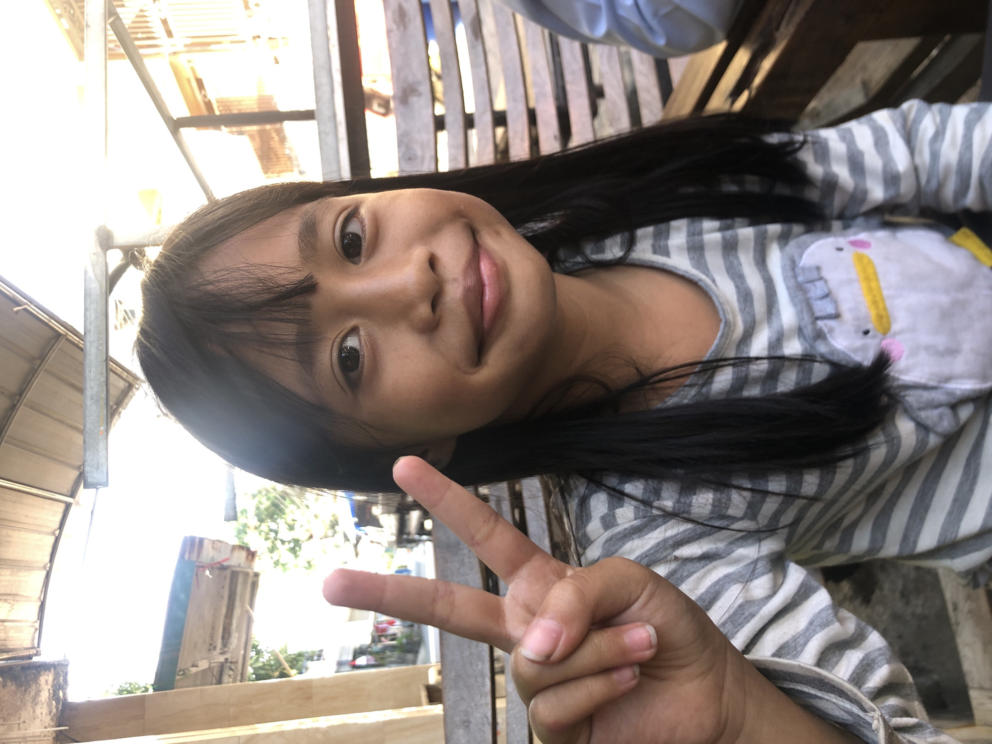 A young girl with long, dark hair and bangs smiles at the camera, holding up her hand in a peace sign. She is wearing a grey and white striped long-sleeved shirt with a cartoon penguin graphic on the front. She is sitting on a wooden bench outdoors, with a corrugated metal roof overhead. The background shows some buildings and trees, suggesting an urban or suburban setting in Surabaya, Indonesia, possibly near a street or market due to the visible structures and vehicles. The lighting indicates it is daytime, with strong sunlight casting shadows and creating a bright, slightly overexposed effect.