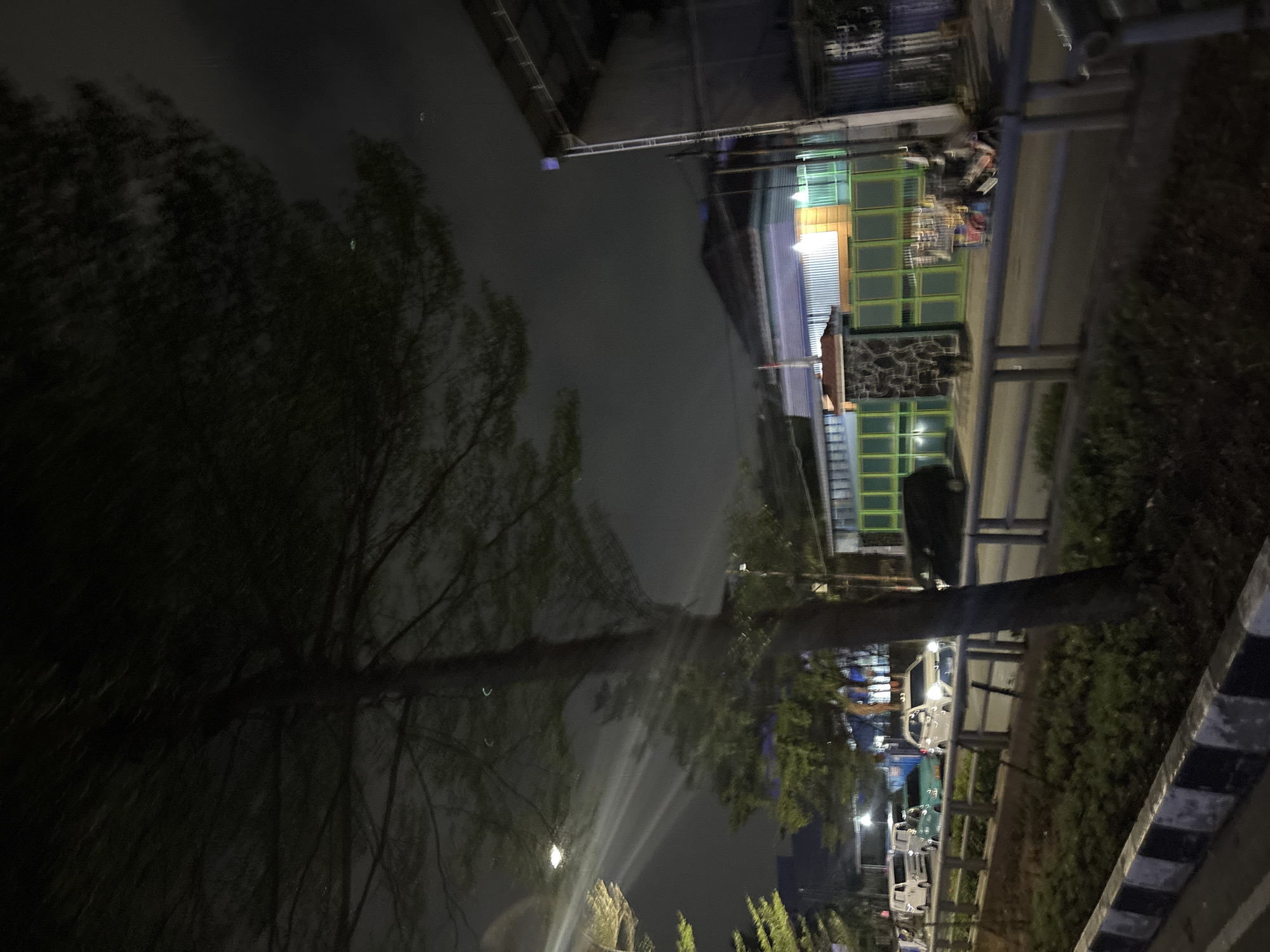 A nighttime scene in North Jakarta, Indonesia features a tree in the foreground with its branches obscuring the sky, which is dark and cloudy. Streetlights cast beams of light through the tree's foliage. In the background, a building with a light blue facade and multi-colored accents is visible, behind a green metal fence. Several vehicles, including white vans, a teal car, a white sedan, and a black SUV, are parked or driving along a road. A guardrail separates the road from a strip of greenery. The scene appears to be a residential or commercial area.