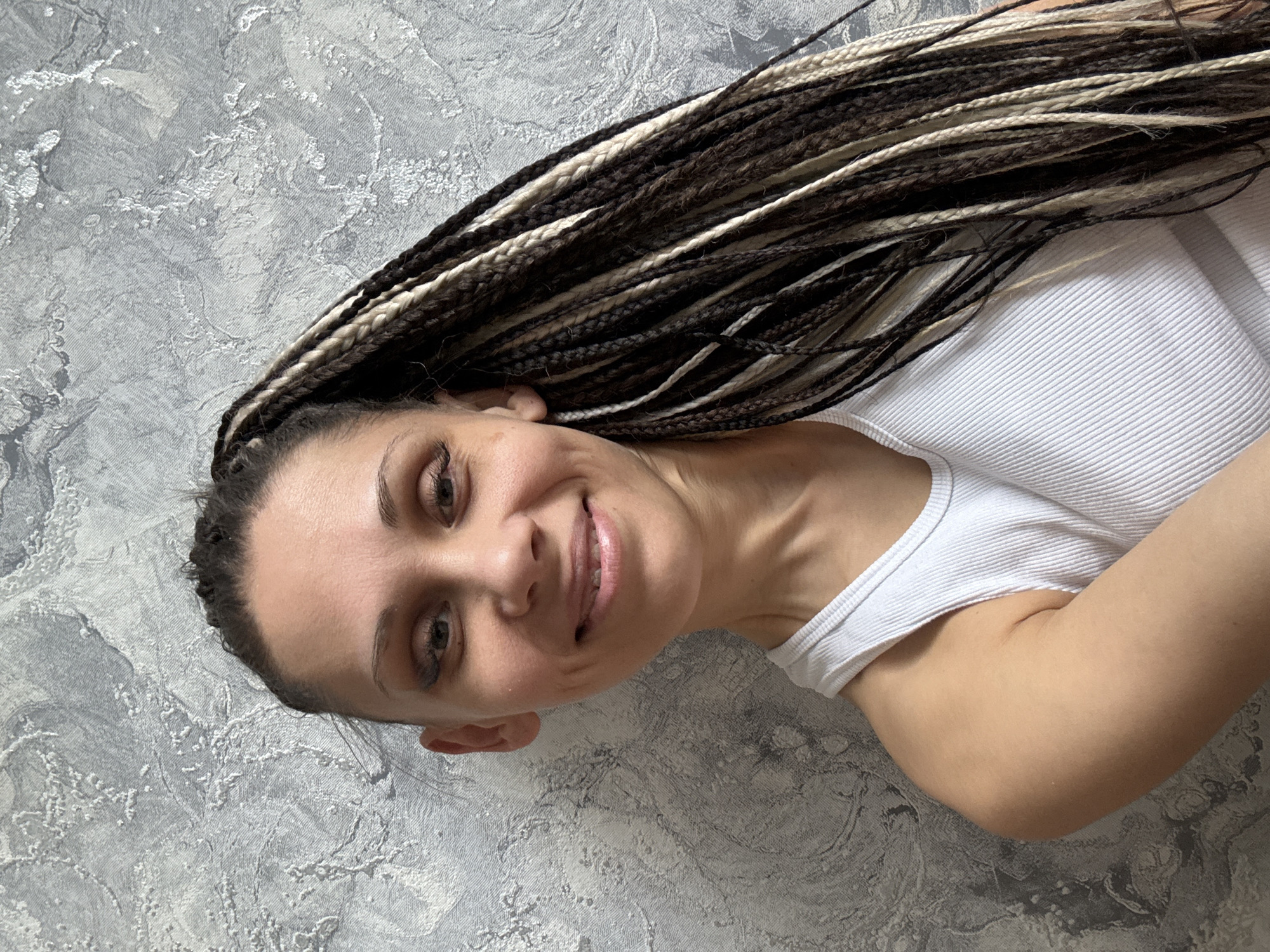 A medium shot depicts a woman with tan skin, brown eyes, and a smiling expression. Her dark brown hair is styled into long braids with interspersed blonde extensions. She is wearing a white ribbed tank top. The background consists of a gray wall with a textured, marble-like pattern. The photo seems to have been taken indoors. The woman's location is unknown.