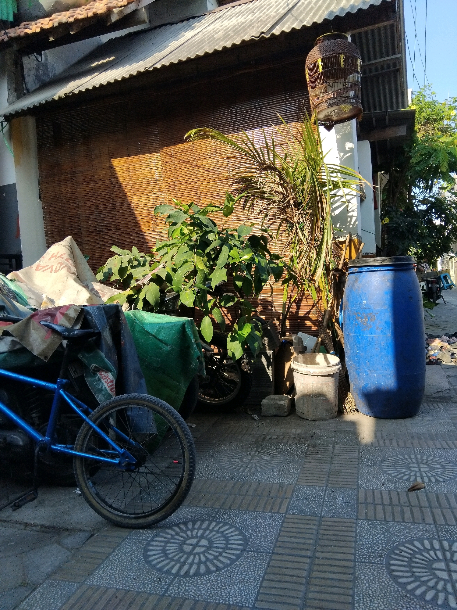 The image depicts an outdoor scene in what appears to be a residential area in Kepuh Kiriman, Indonesia. In the foreground, a blue bicycle is parked on a patterned tiled walkway. The bicycle is partially covered by a large, light-colored tarp. Behind the bicycle, several objects are visible, including what looks like a large green tarp draped over other items, and a tall blue plastic barrel. Further back, a house with a corrugated metal roof and bamboo blinds is visible. A large, ornate birdcage hangs from the roof's overhang. Lush green foliage surrounds the birdcage and the house. Sunlight casts shadows across the scene, indicating it is daytime. The overall impression is one of an outdoor storage area or a less-maintained section of a property. No people are visible in the frame.