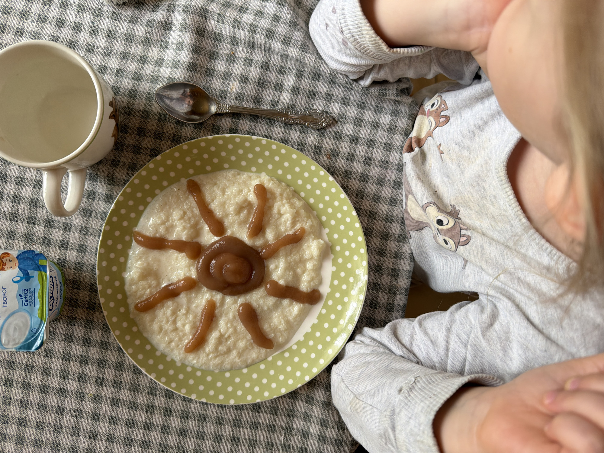 The image shows a child at a table eating. A plate of porridge decorated to resemble a sun is visible, along with a cup, a spoon, and a container of Tvorog cheese. The child is wearing a long-sleeved shirt with cartoon characters on it. The table is covered with a gray and white checkered tablecloth. The location is implied to be Khimki, Russia, based on the labeling of the Tvorog cheese container.