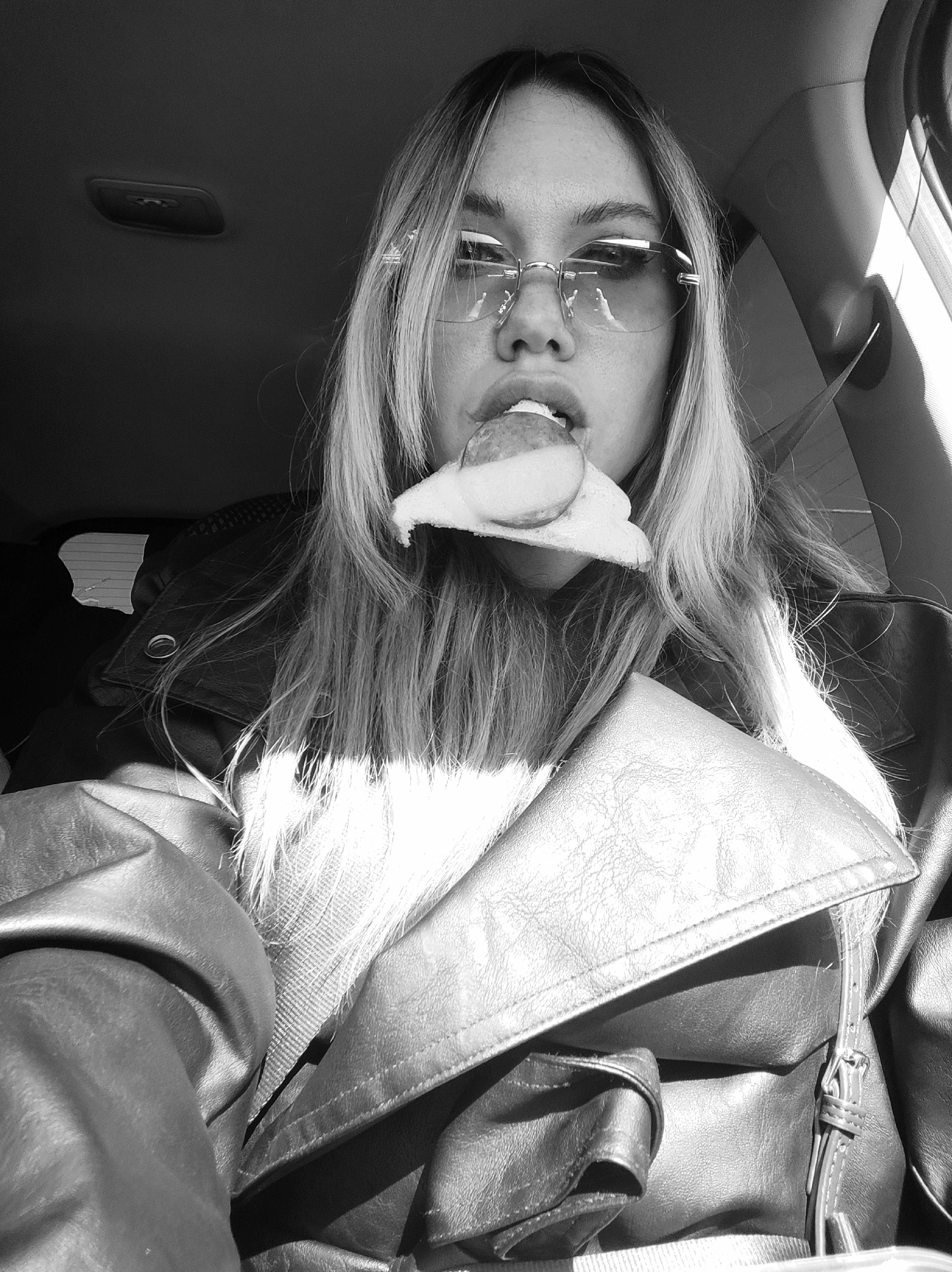 The image depicts a woman in the interior of a car. She is wearing sunglasses and a leather jacket. She has long hair. She is holding a slice of food in her mouth. A seatbelt is visible across her chest. The location is indeterminable from the image. The city of Izhevsk, Russia, cannot be confirmed as the location based on the visual information provided.