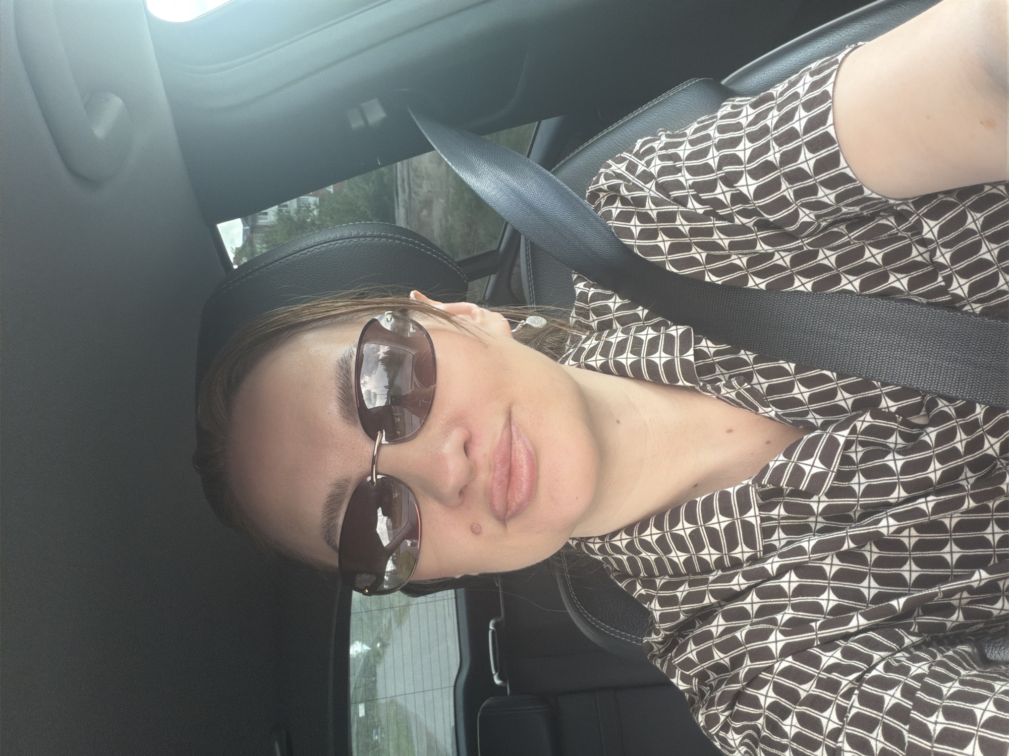 A woman, appearing to be of Slavic descent, is captured in a selfie taken from the driver's seat of a car. She is wearing sunglasses and a brown and white patterned shirt, with a seatbelt fastened across her chest. The background suggests an outdoor setting, possibly a street or residential area, with glimpses of buildings and trees visible through the car windows. The lighting indicates it is daytime, likely sunny. The image appears to have been taken in Mezhevoy, Russia.