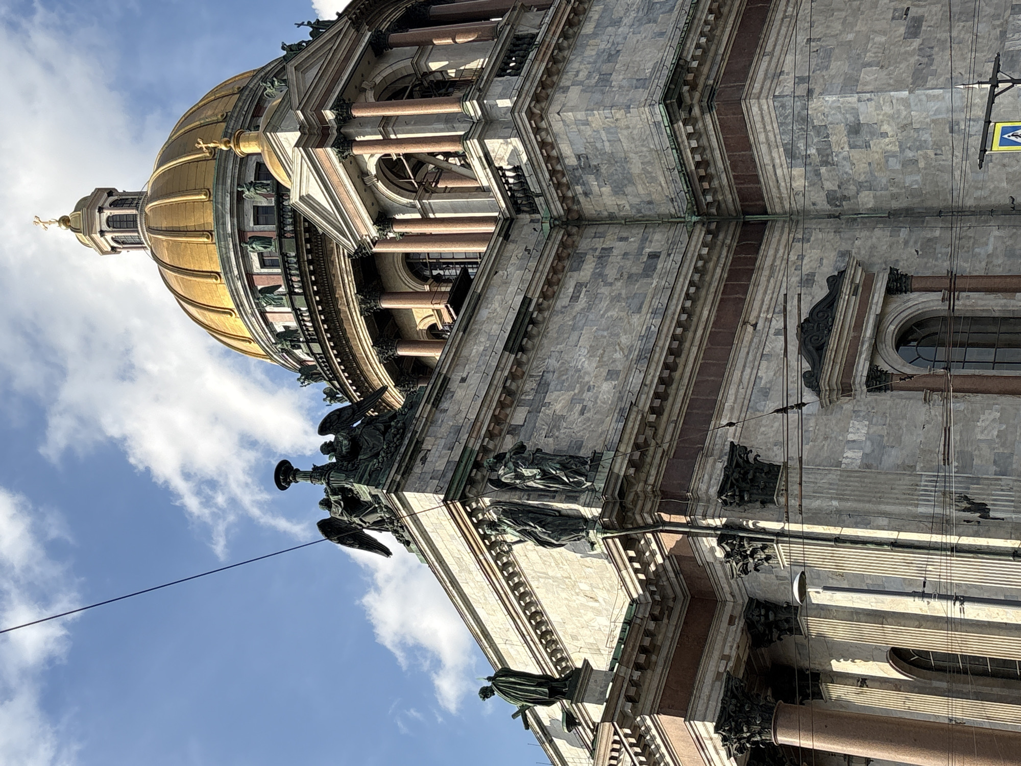 The image depicts a low-angle shot of Saint Isaac's Cathedral in Saint Petersburg, Russia. The angle emphasizes the height and architectural details of the building, showcasing the dome, columns, and various statues. The sky is visible in the background, with some clouds. There are no people visible in the image. The photo presents the cathedral as a massive and ornate structure.