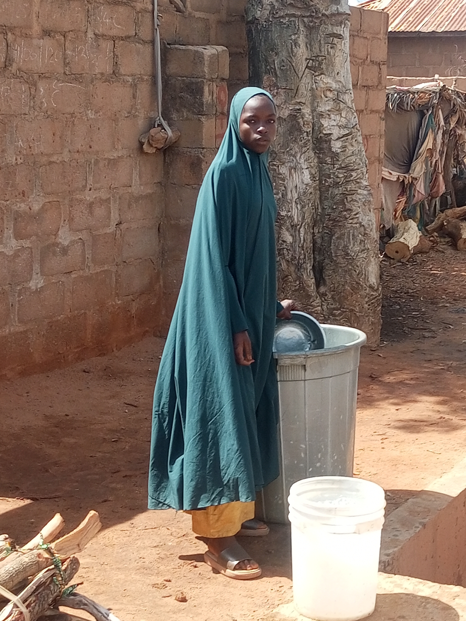 This image depicts a young person standing outdoors in a rural or semi-rural setting in Tempure, Nigeria, under bright daylight with strong shadows. The individual wears a teal-green hijab and a long, flowing outer garment, revealing yellow trousers and sandals underneath. They are positioned next to a large, light grey plastic bin, holding a metal plate over its opening, possibly in the process of washing or handling items.
To the left of the person is a rustic brick wall, upon which faint handwritten numbers and letters are visible, including "11/00/120 12+" and other scribbles. A thin rope, from which a stone is tied and hanging, is attached to the wall. Behind the person and the bin stands a large tree with a rough, textured trunk. The ground is bare earth.
In the lower left foreground, a bundle of firewood, bound together, rests on the ground. A smaller, white plastic bucket is situated on the ground to the right of the larger bin. In the distant background, hints of simple structures, possibly with corrugated metal roofing, can be discerned. The person's expression is neutral, as they look directly towards the camera.