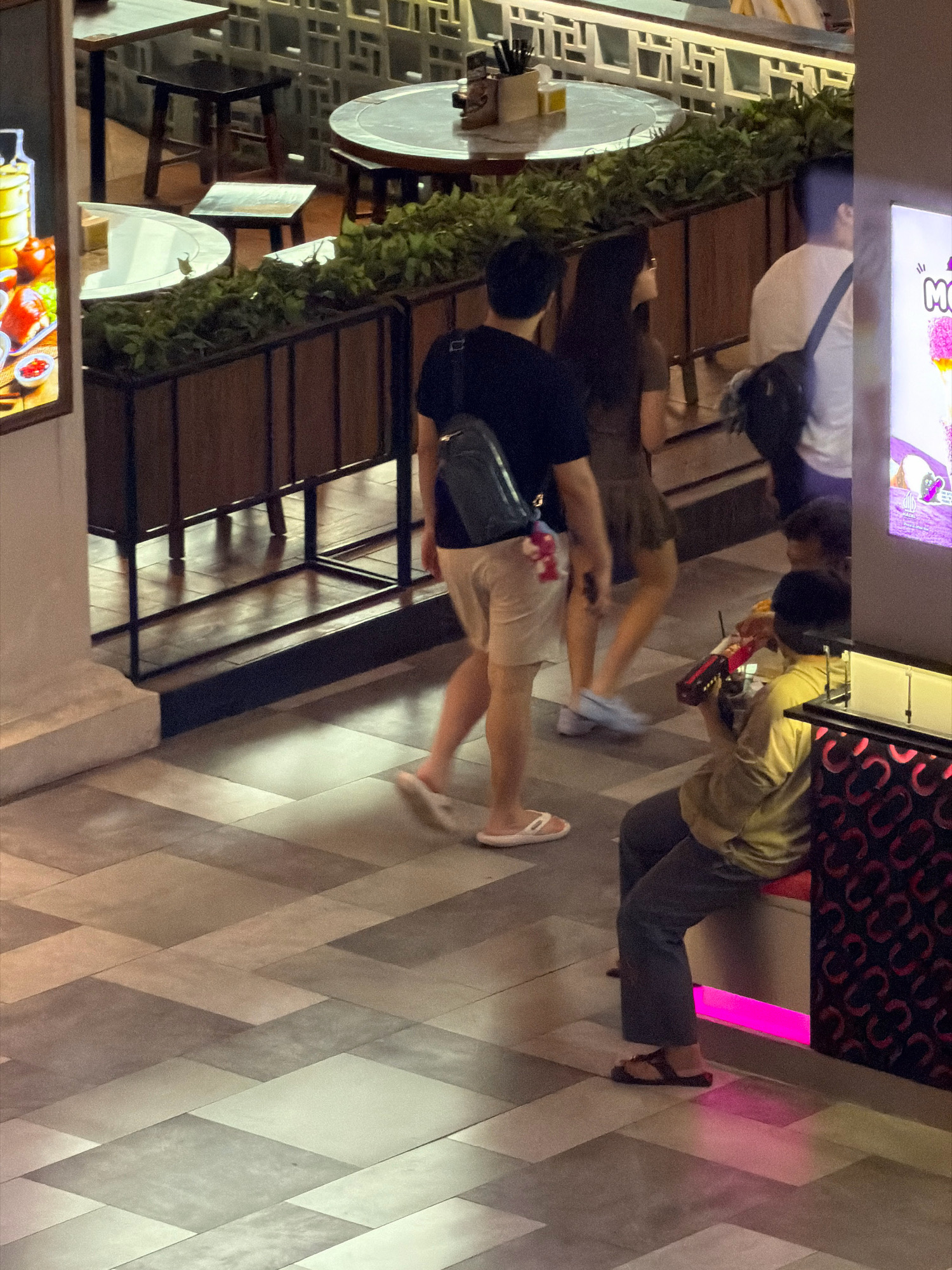 The image shows an indoor scene with people walking and sitting on a tiled floor. In the foreground, a person wearing a light brown shirt and dark pants is sitting on a red bench, holding a red toy gun. To their left, a person in a black t-shirt and light-colored shorts walks away from the camera, carrying a dark bag over their shoulder. Behind them, a person in a brown dress is walking, followed by another person in a white t-shirt carrying a black backpack. In the background, there are tables and chairs, suggesting a dining or seating area, with decorative greenery. The lighting is dim, with some areas illuminated by pink neon lights.