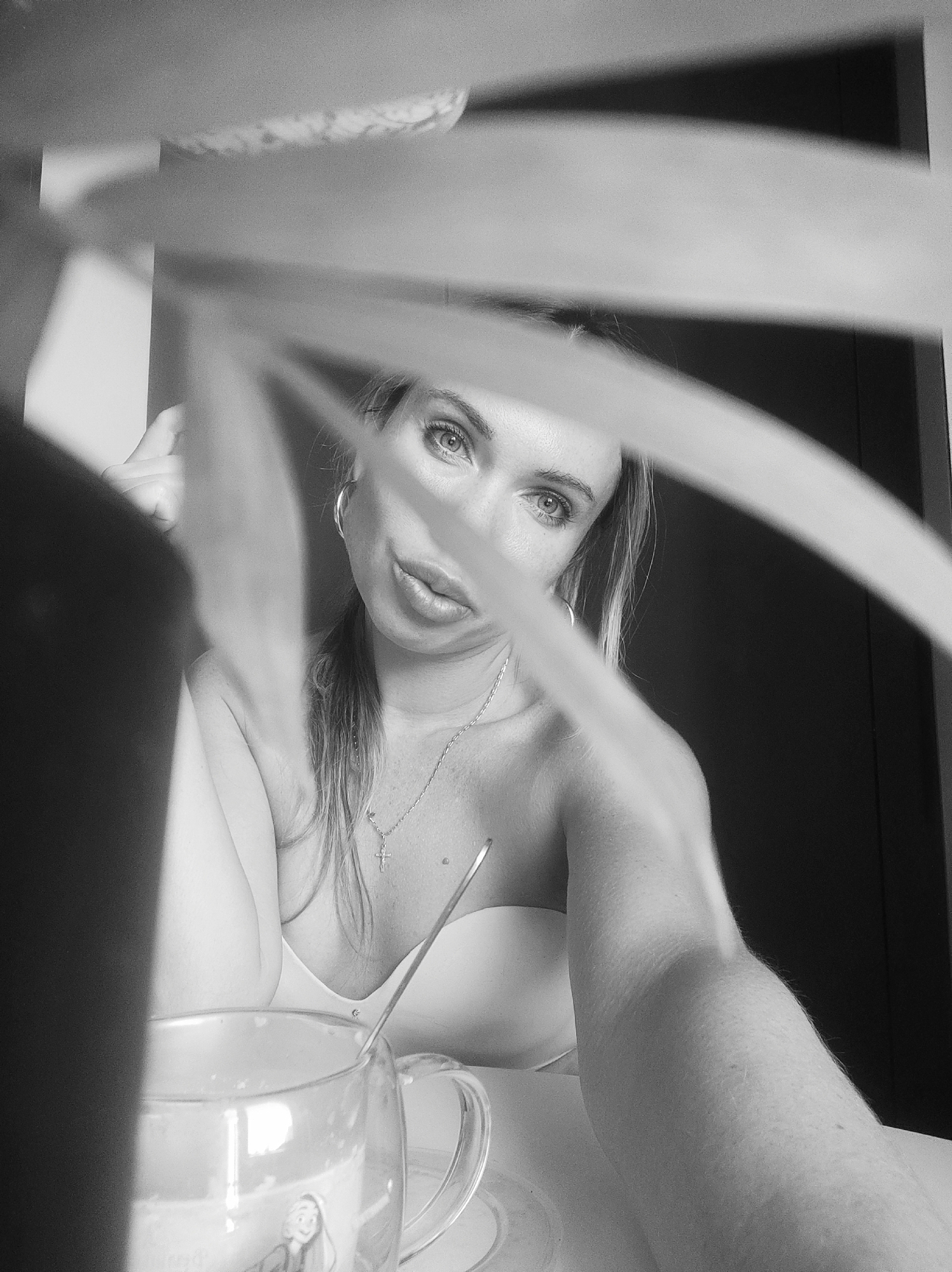 A black and white portrait shows a fair-skinned woman with light eyes, wearing hoop earrings and a delicate cross necklace. She is partially obscured by the leaves of a plant in the foreground. A clear glass mug with a spoon inside sits on a table. The background is a dark, solid color. There is no visible information to indicate this photo was taken in Votkinsk, Russia.