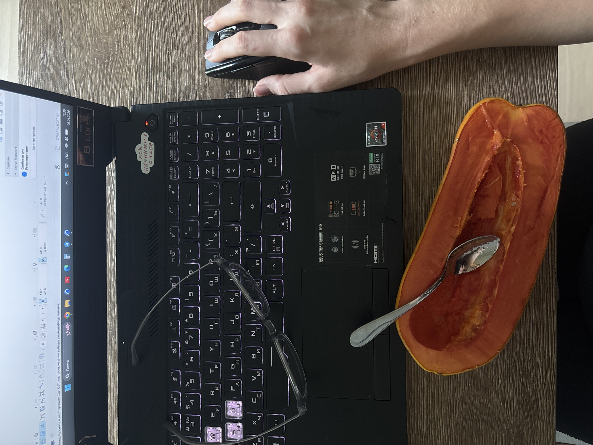 A top-down view shows a laptop on a wood-patterned surface with a papaya half containing a spoon. A hand operates a black computer mouse to the right of the laptop. On the laptop's keyboard are a pair of eyeglasses. The laptop screen displays a design software interface. The laptop has branding stickers. The location is not discernable from the given image.