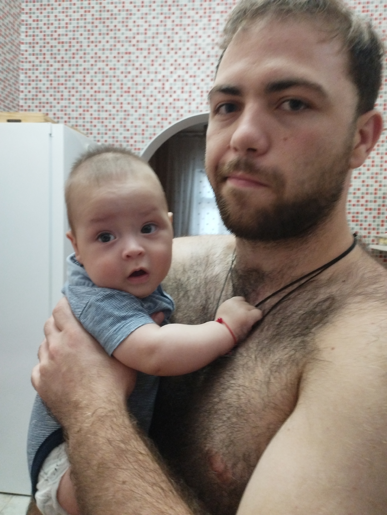 The image shows a man holding a baby. The man is shirtless and has a lot of chest hair. The baby is wearing a blue and white striped shirt and a diaper. The background appears to be a kitchen with red and white tiled walls and a white cabinet. The overall scene is a candid snapshot, likely taken in a domestic setting. No specific details regarding the city of Fălești, Moldova are visible within the image itself.