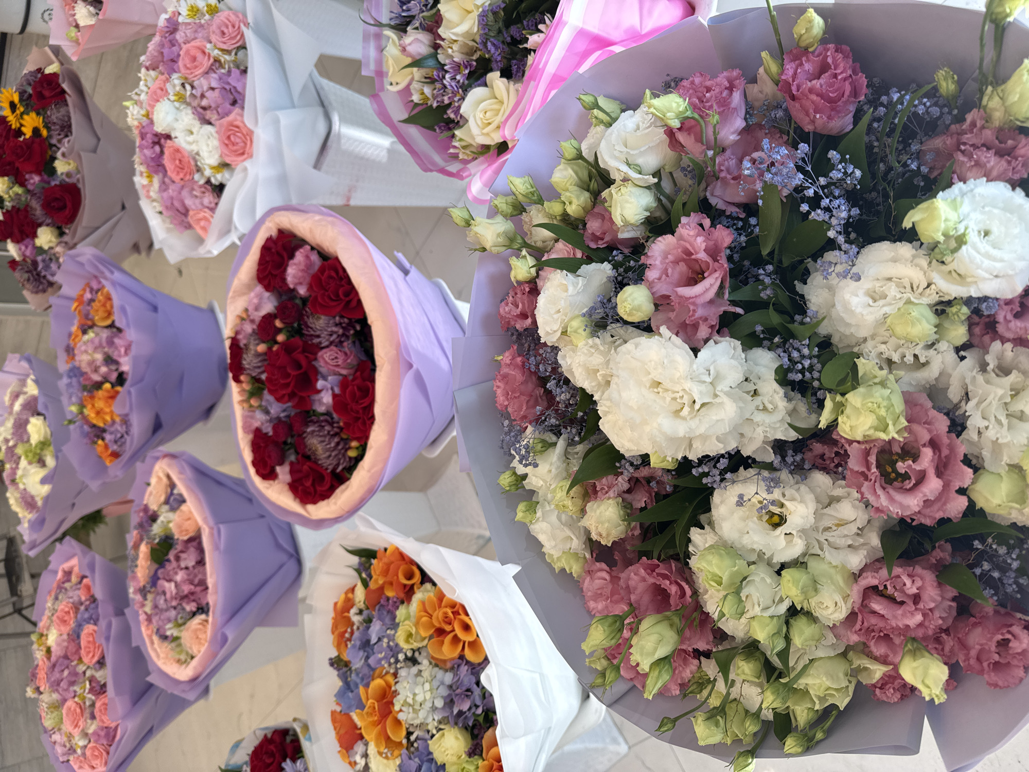 The image displays a close-up, eye-level, outdoor shot of numerous bouquets of flowers arranged for sale. The flowers are predominantly in shades of pink, purple, white, red, and orange, with greenery visible throughout. The bouquets are wrapped in various colored papers, including lavender, pink, and white. The scene suggests a floral shop or market setting. There are no people or specific city landmarks visible, therefore it is not possible to determine if this is in Baku, Azerbaijan.