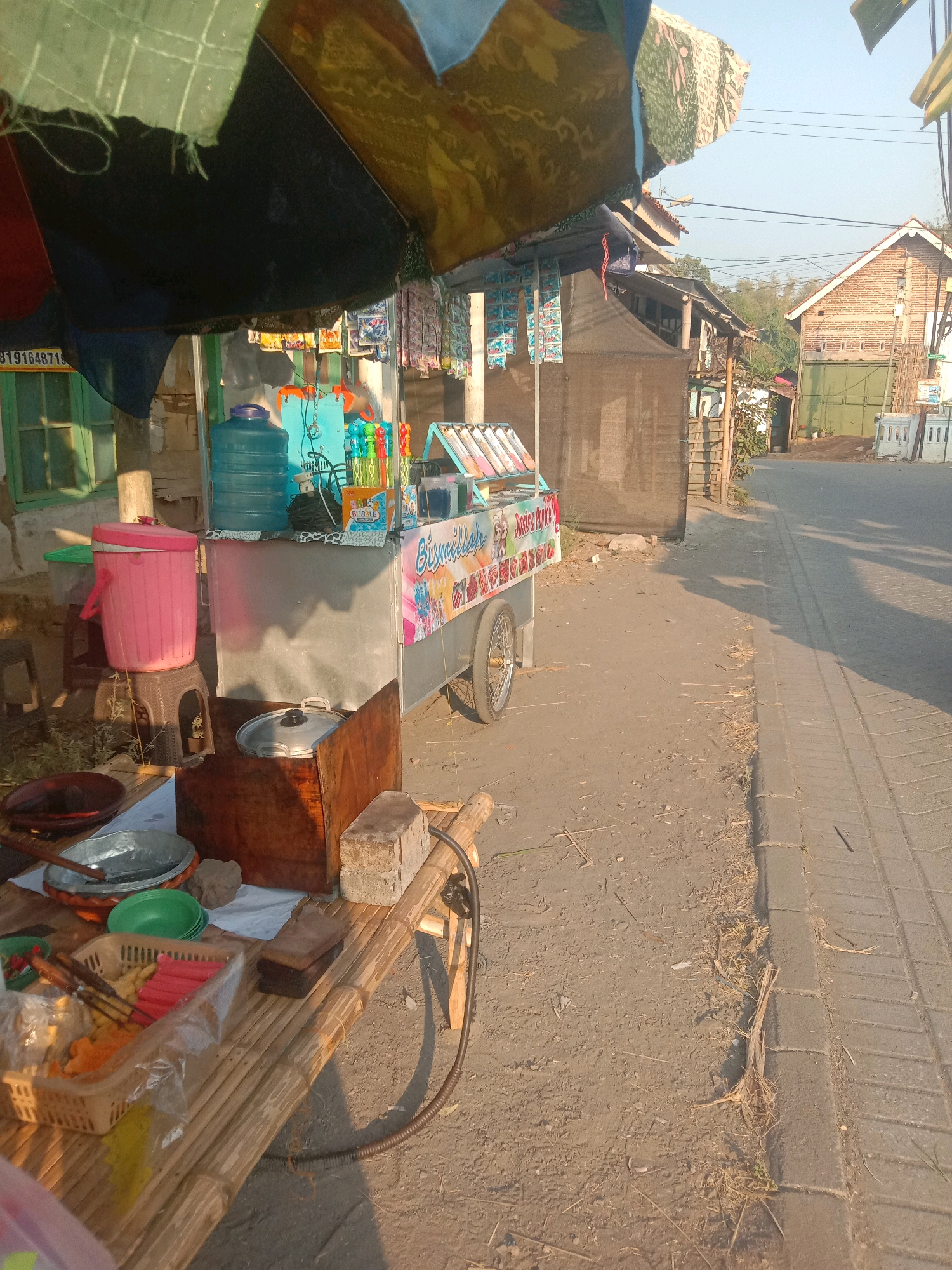 A street-side food cart is visible in Selotambak, Indonesia, bathed in the warm light of what appears to be late afternoon sun. The cart itself is colorful, with a sign featuring cartoon characters and text in Indonesian. It is stocked with various food items and drinks, including a large blue water jug, bottled drinks, and a basket filled with what appear to be snacks like sausage and fried items. Next to the cart, a makeshift bamboo table holds cooking utensils, a pot with a lid, and a pink trash can on a stool. Behind the cart, there are several small buildings and a brick structure. The street itself is paved on the right side, with a dirt shoulder where the cart and table are situated. The environment suggests a simple, local market or roadside eatery.