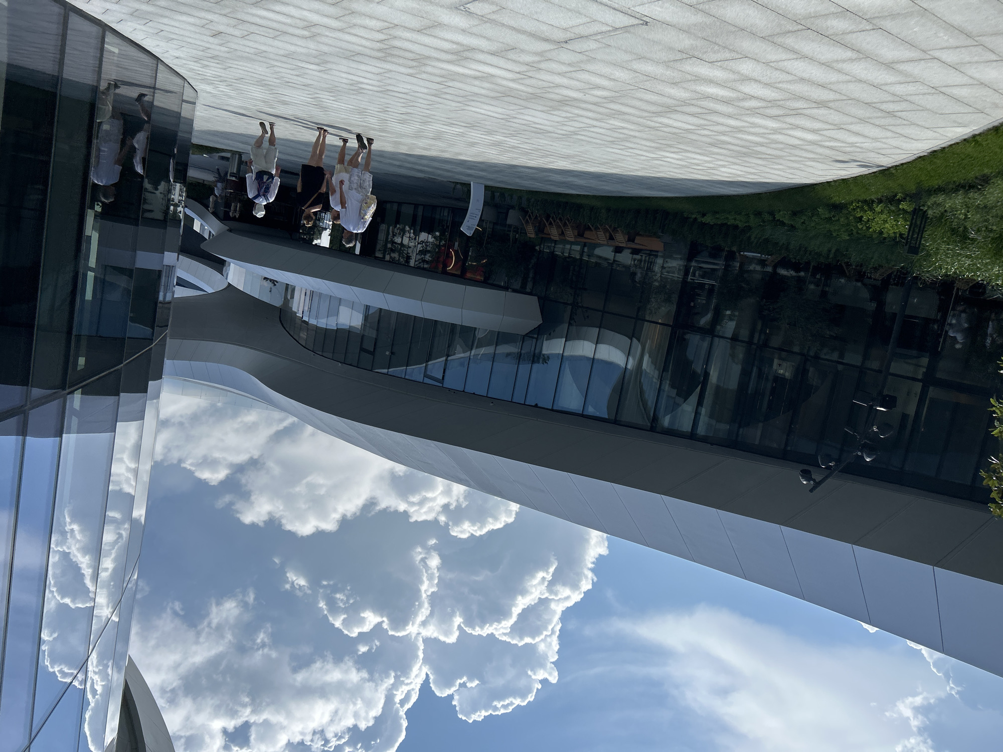 This image depicts the exterior of the Gelendzhik Congress Center, located in Gelendzhik, Russia. The architecture features a modern design with a curved, white facade and extensive glass paneling that reflects the sky and surrounding environment. In the foreground, a paved walkway made of light-colored rectangular stones curves towards the building. Several individuals are walking on the walkway, appearing to be tourists or visitors. One group of three is in the middle ground, walking away from the camera. Another individual is seen walking further into the distance, and a person is reflected in the glass of the building. Lush green vegetation, including grass and shrubs, borders the walkway. The sky above is a clear blue with large, fluffy white clouds, suggesting a bright, sunny day. The overall scene conveys a sense of modern urban development and leisure.