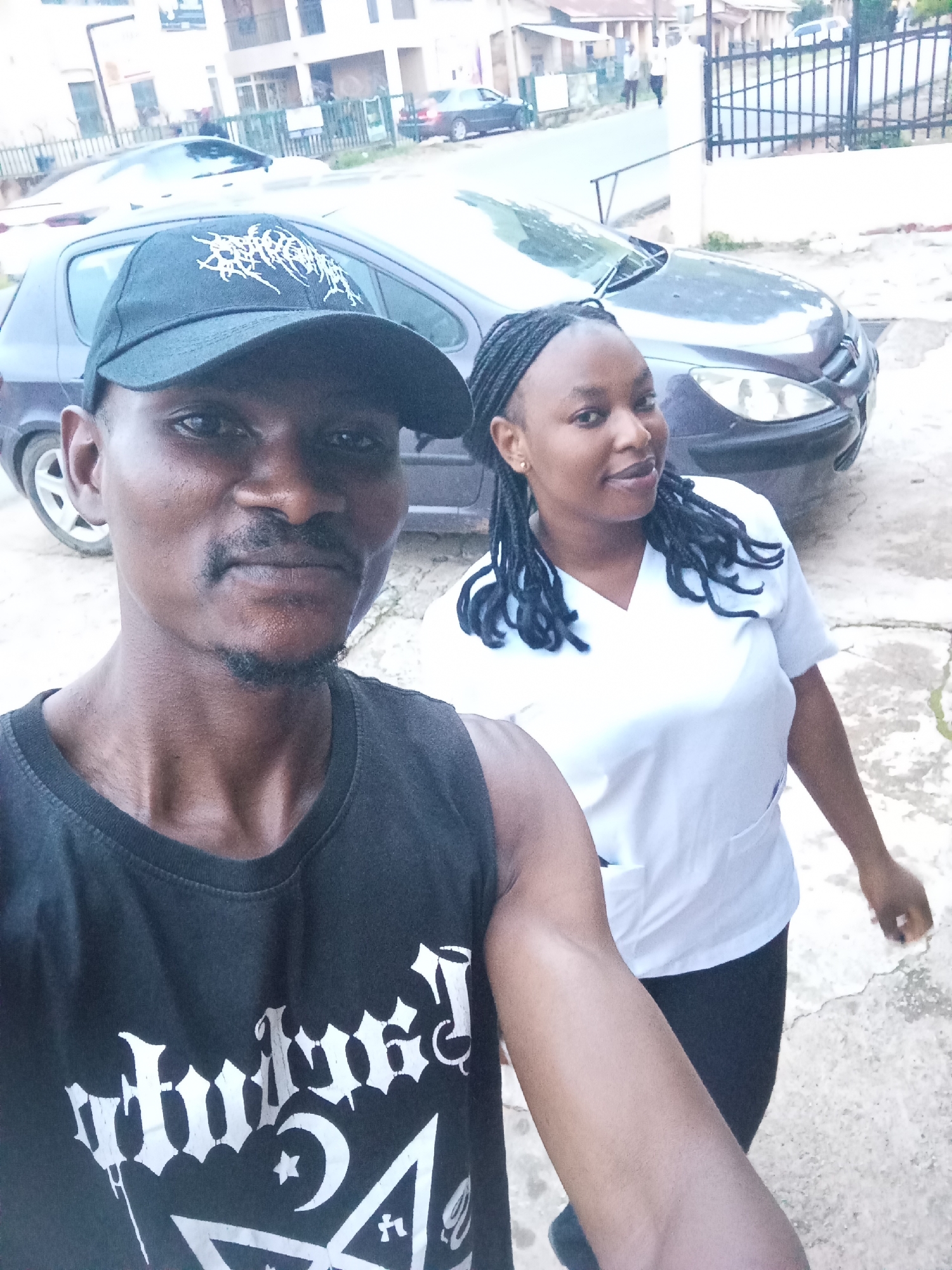 A medium shot selfie depicts two individuals, a man and a woman, standing outdoors. The man, positioned in the foreground, wears a black sleeveless shirt with white gothic-style lettering and a black baseball cap. He has dark skin, a mustache, and a goatee. The woman, slightly behind and to the right of the man, wears a white V-neck scrub top and black leggings. She has dark skin, braided hair, and is looking towards the camera with a slight smile. Behind them, a dark-colored hatchback car is visible, parked on what appears to be a paved surface. In the background, buildings and a street with a metal fence can be seen. The overall scene suggests a casual outdoor setting, possibly a residential or commercial area in Jos, Nigeria, given the context. The lighting suggests it is daytime.