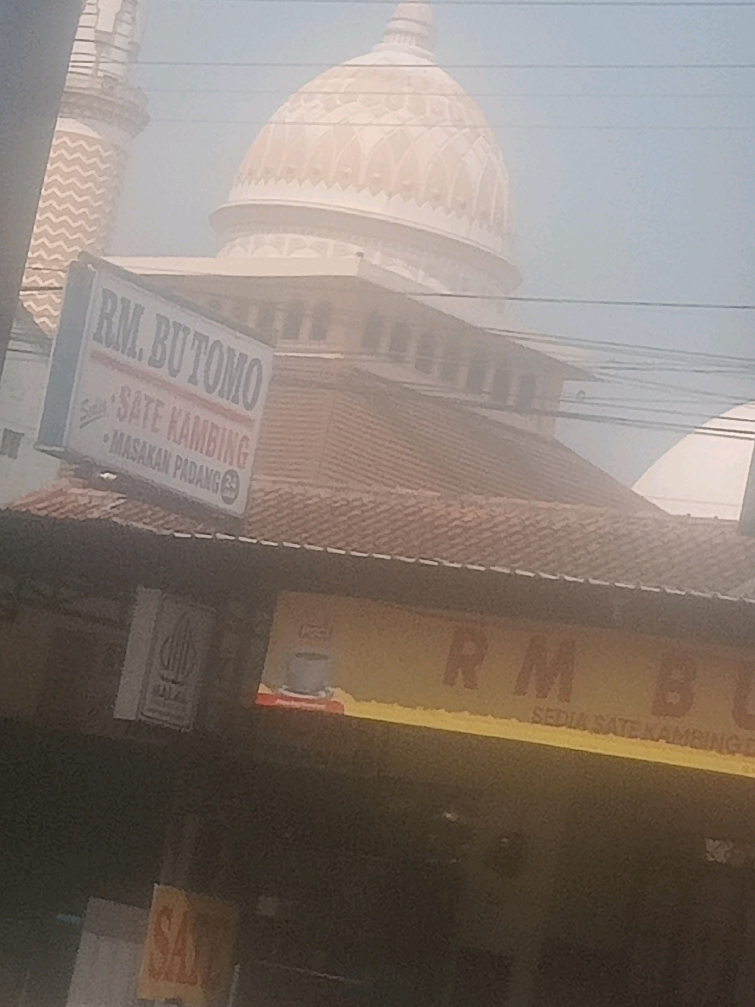 The image shows a street scene in Lebaksiu, Indonesia. In the background, there is a large mosque with a dome and minarets. In the foreground, there is a building with a sign that reads "RM. BUTOMO SATE KAMBING - MASAKAN PADANG". Another sign below it reads "RM B SEDIA SATE KAMBING D". A smaller sign with a halal logo is also visible on the building. The scene appears to be outdoors, with some atmospheric haze. There are no people clearly visible in the image.