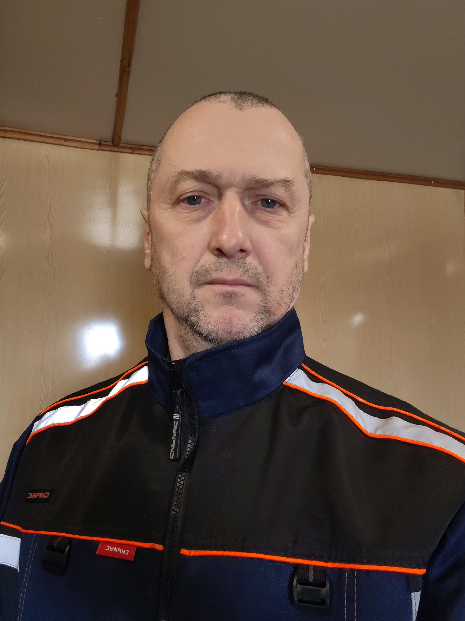 A middle-aged man with fair skin, short salt-and-pepper hair, and a trimmed beard is facing the camera. He is wearing a work jacket with black and blue sections, orange piping, and reflective white stripes. The jacket has a zipper and logos/text that appear to read "CNBNAC." The background is a plain wall, possibly wood paneling. The image was taken in a location within the city of Unknown, Unknown.