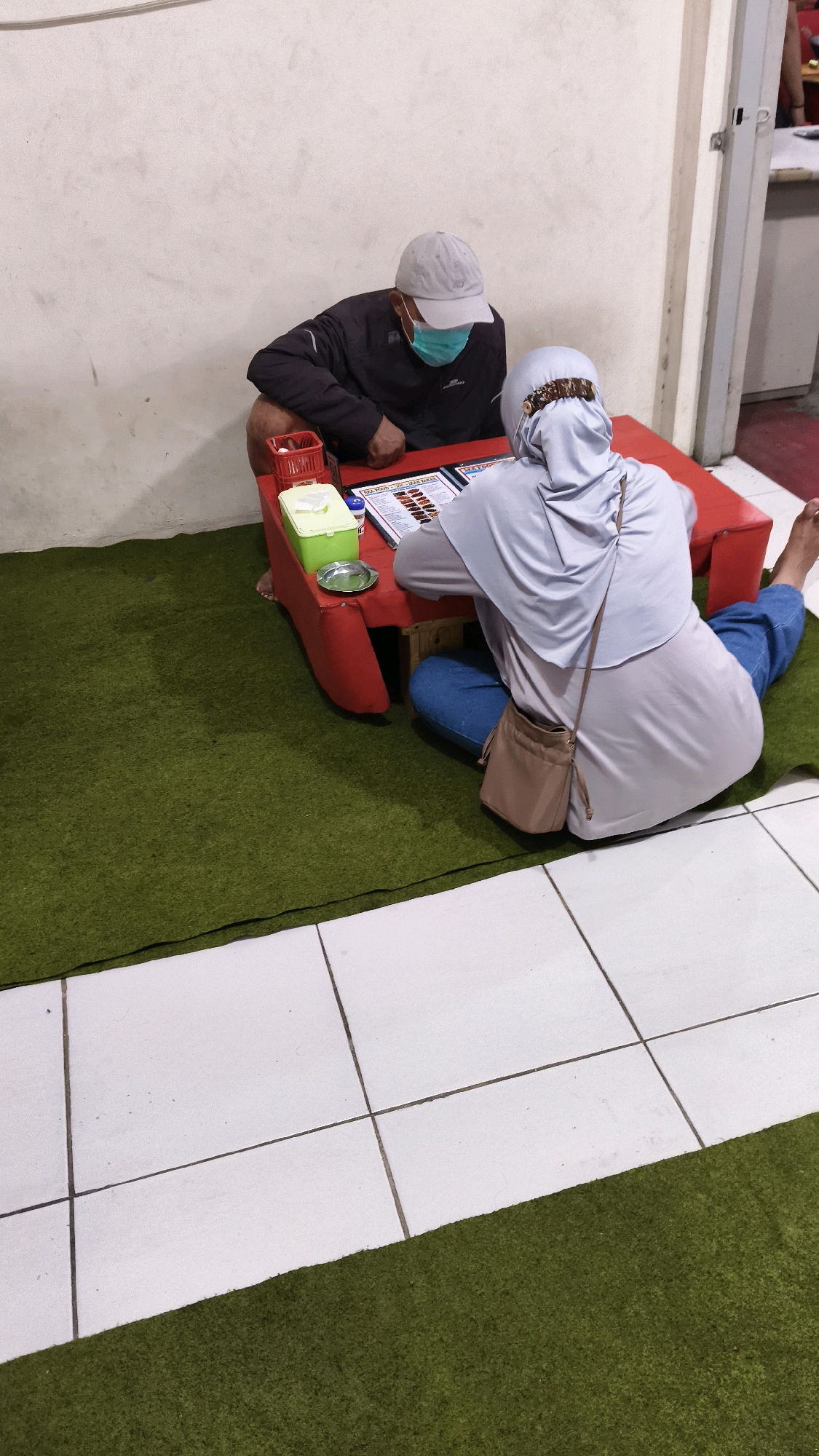 Two individuals, a man and a woman, are seated on a green carpeted floor, facing a red table. The man, wearing a grey baseball cap and a light blue face mask, is leaning forward, looking down at the table. A dark jacket is visible over his shoulders. The woman, who is wearing a light blue hijab and a light blue top, is also looking at the table, facing away from the camera. She has a small tan crossbody bag slung over her shoulder. On the table, a menu is open, and a green container with tissues and a small metal dish are present. The setting appears to be an indoor establishment with a white wall behind the individuals and tiled flooring visible in the foreground. The image was taken in the city of Karangsatria, Indonesia.