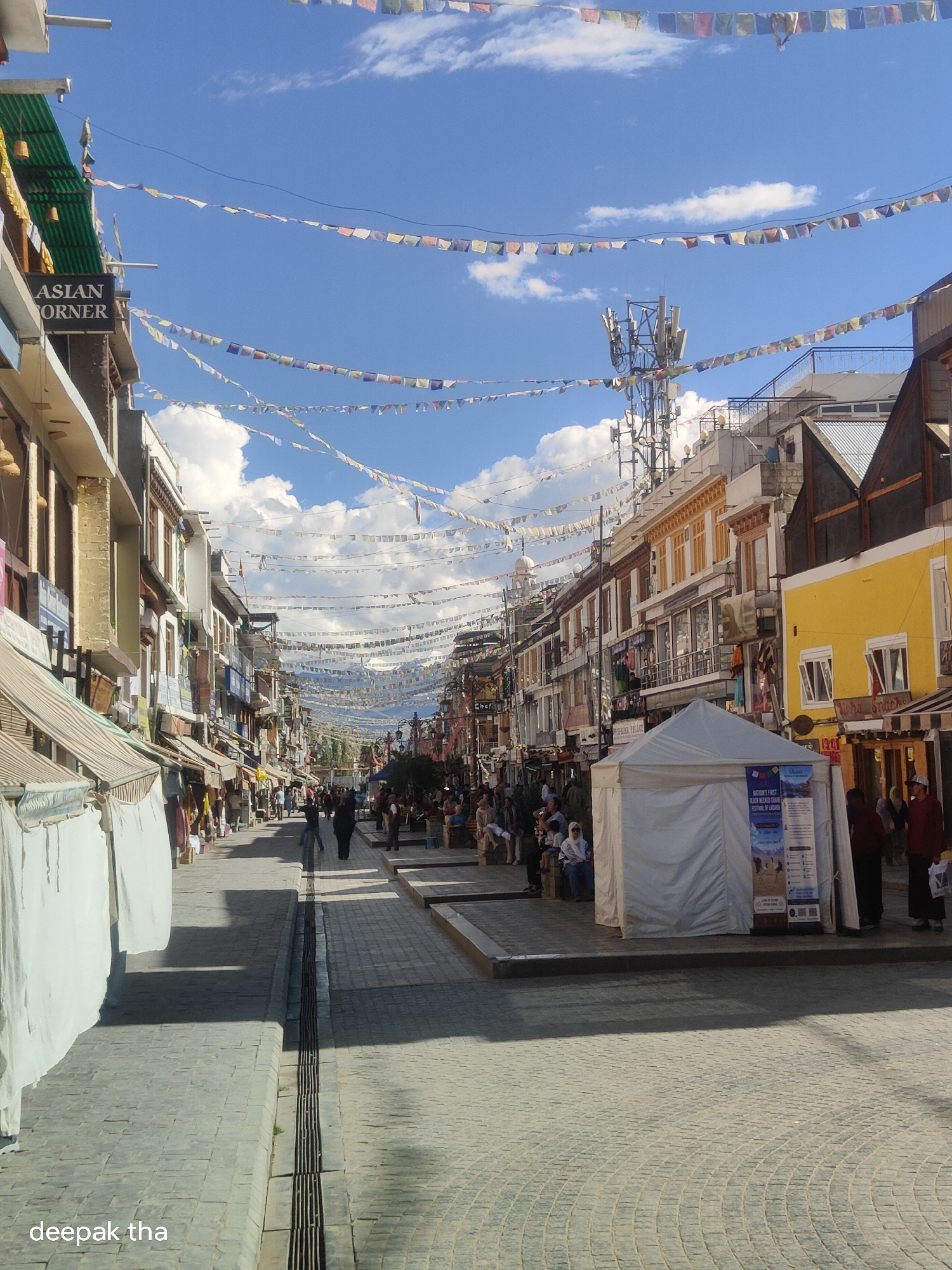 A street in Leh, India, is lined with shops and adorned with prayer flags. A white tent is set up on the right side of the street, with a poster displayed on it. People are walking along the street and sitting at outdoor tables. The sky is blue with some clouds.
