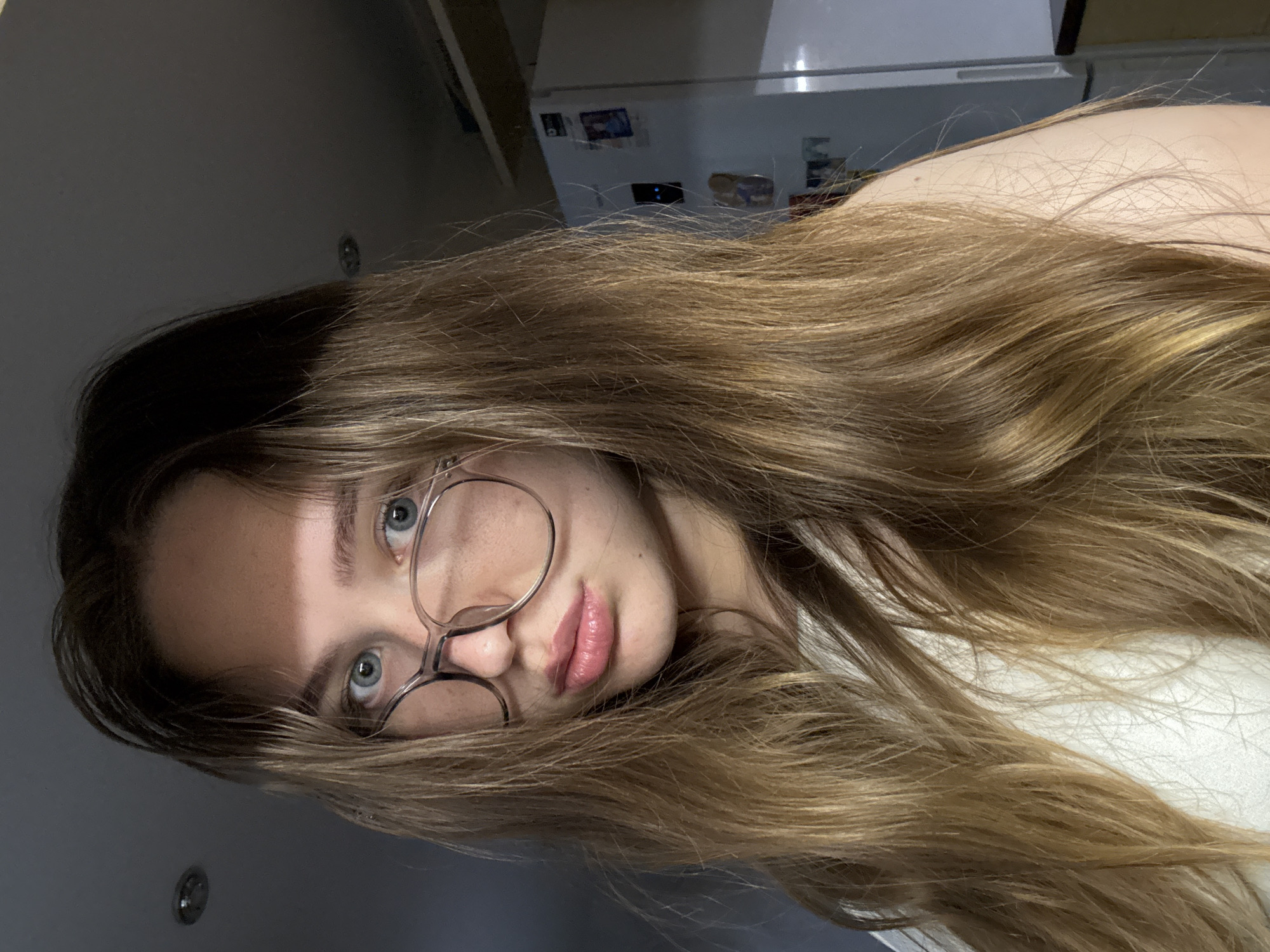A young woman with long, blonde hair and glasses is posing for a close-up selfie indoors. She is wearing a white top. The background shows a refrigerator and a kitchen cabinet, suggesting the location is a domestic kitchen. The lighting is dim with a shadow falling across her forehead and eyes. There is no indication of the specific location within the city of Chaikovsky, Russia.