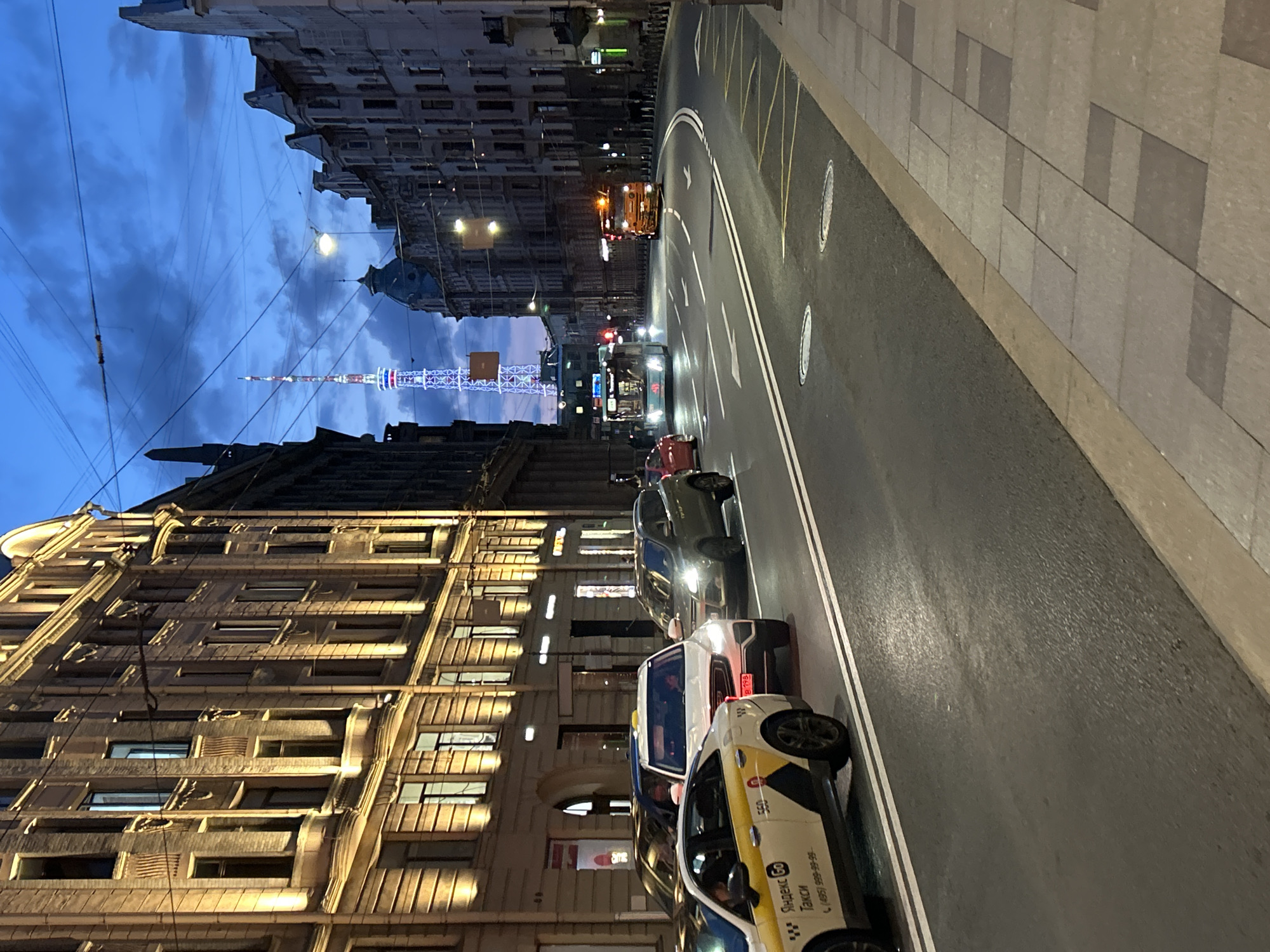 In a city scene at dusk, a wide asphalt street curves to the right, bordered by sidewalks. On the left, a row of tall, ornate buildings with illuminated windows and decorative facades lines the street. A yellow taxi with "Яндекс Go" and "Такси" branding is parked in the foreground, partially obscuring other vehicles. Further down the street, traffic is visible, including a light blue bus with illuminated signs and an orange truck with flashing lights. In the distance, a tall, slender tower with intricate lighting rises against the twilight sky, which is filled with dramatic clouds. The street is marked with white lane lines and yellow markings on the edge. Several manhole covers are visible on the road surface. The overall atmosphere suggests a busy urban environment at the end of the day.