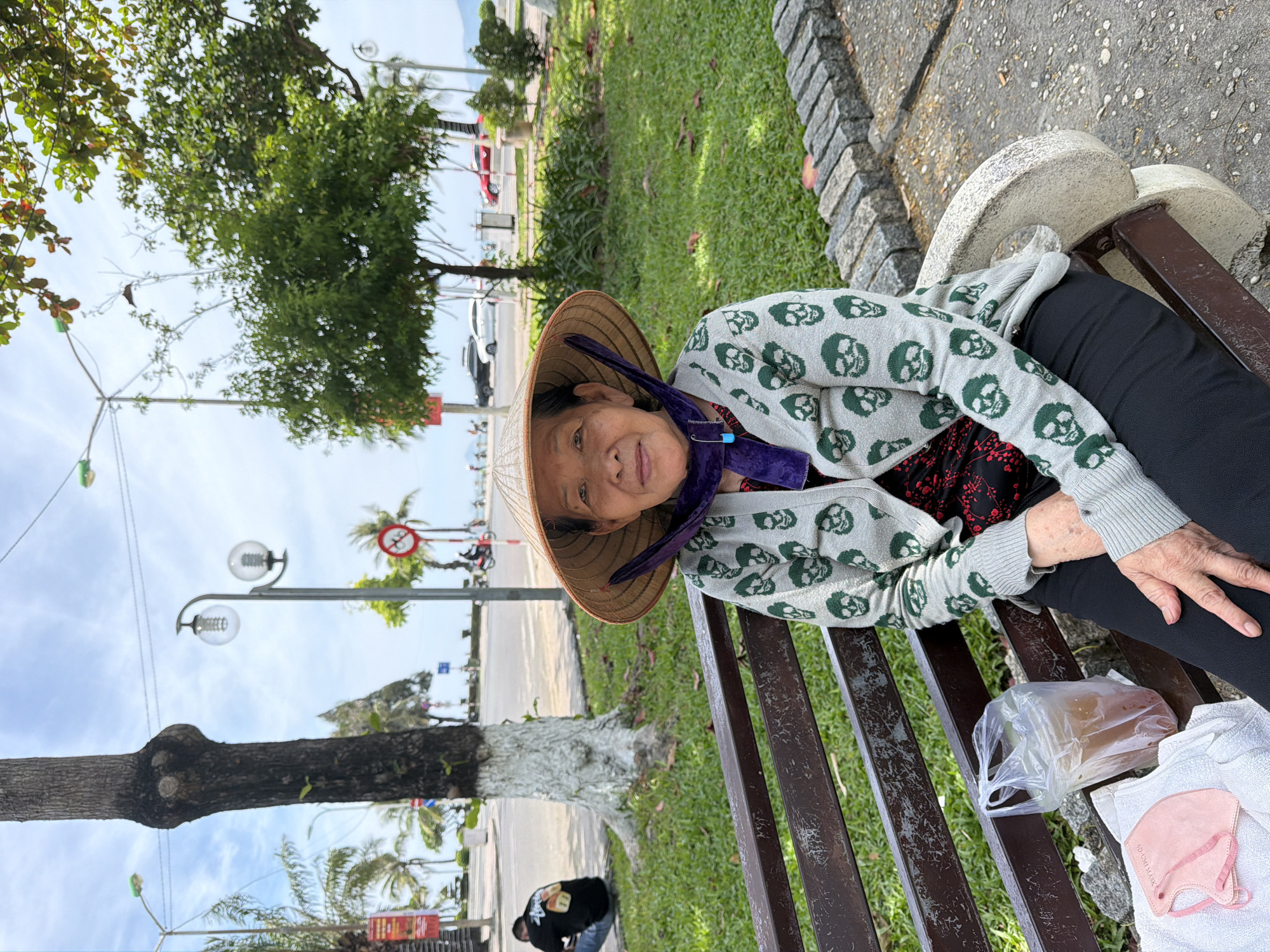 In Nha Trang Ward, Vietnam, an elderly Vietnamese woman wearing a conical hat and a grey cardigan with a skull pattern is seated on a park bench. She is looking at the camera and smiling. In front of her, on the bench, are a plastic bag with a drink and a pink face mask. The bench is made of wood and stone. Behind her, a street is visible with cars and palm trees. The sky is cloudy.