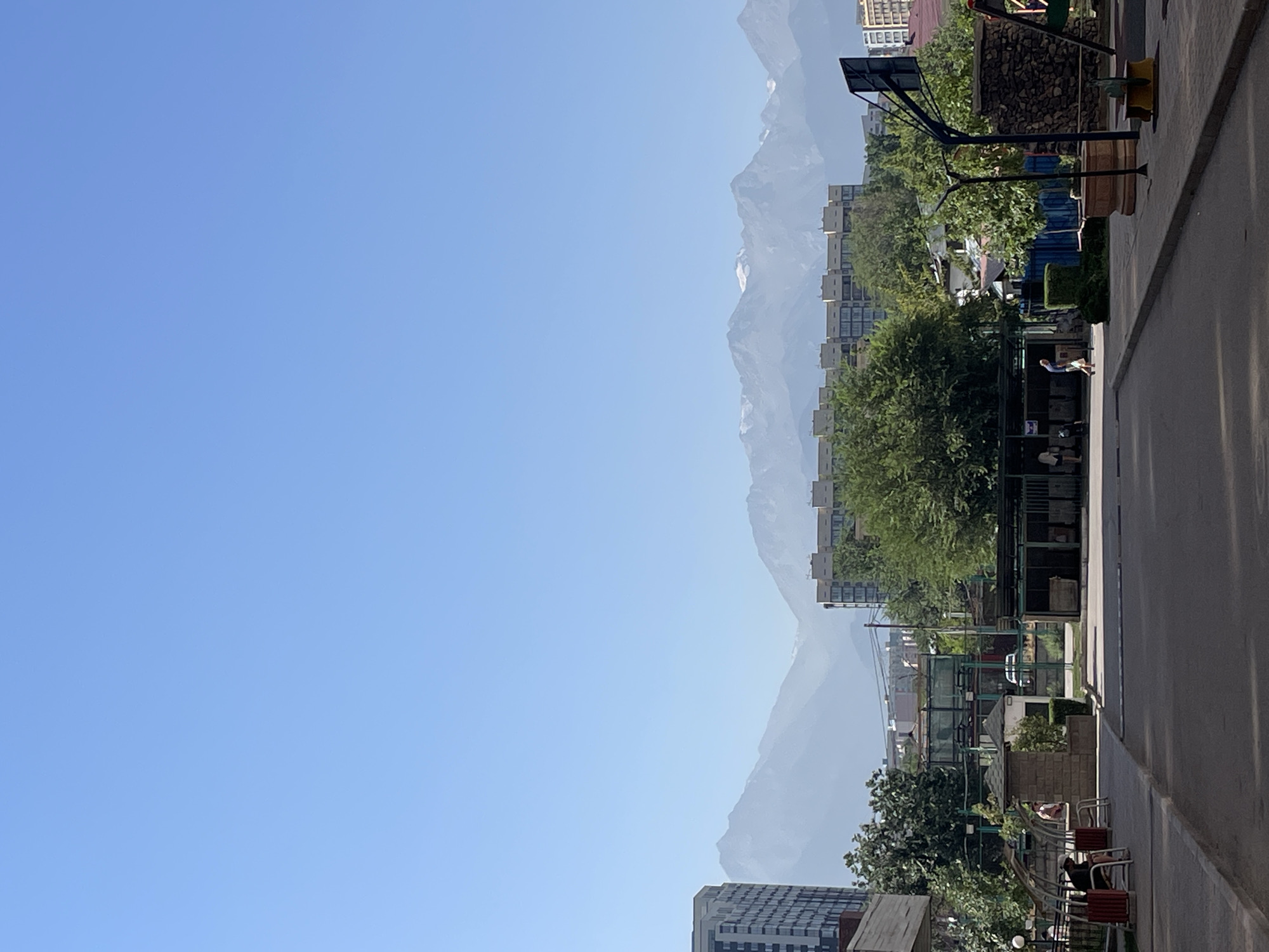 The image depicts a street scene in Almaty, Kazakhstan, with the majestic Zailiysky Alatau mountain range visible in the background under a clear blue sky. In the foreground, a paved street stretches towards the mountains, flanked by sidewalks. To the left, a residential building with a modern facade is visible, along with park benches and trees. On the right, a basketball hoop and net are set up, and a playground with colorful equipment can be seen. Several people are present in the scene, some walking or standing, while others are seated on benches. A covered outdoor seating area with tables and chairs is also visible. The overall impression is that of a residential neighborhood with recreational facilities, set against a stunning natural backdrop.