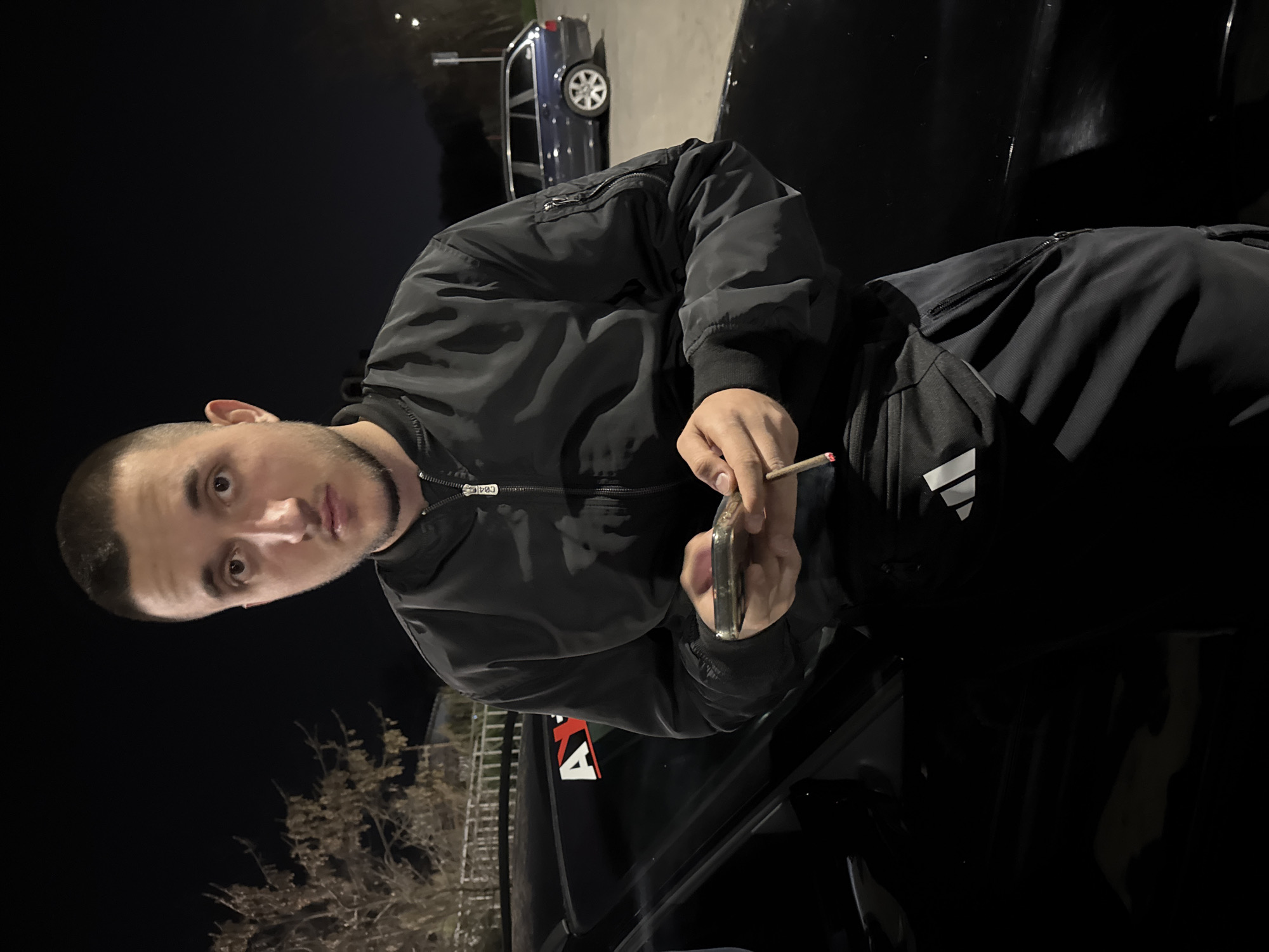 At night in Topoli, Bulgaria, a young man with short hair stands leaning on a black car, holding a lit cigarette and a phone. A blue car is parked in the background. The environment is lit by streetlights.