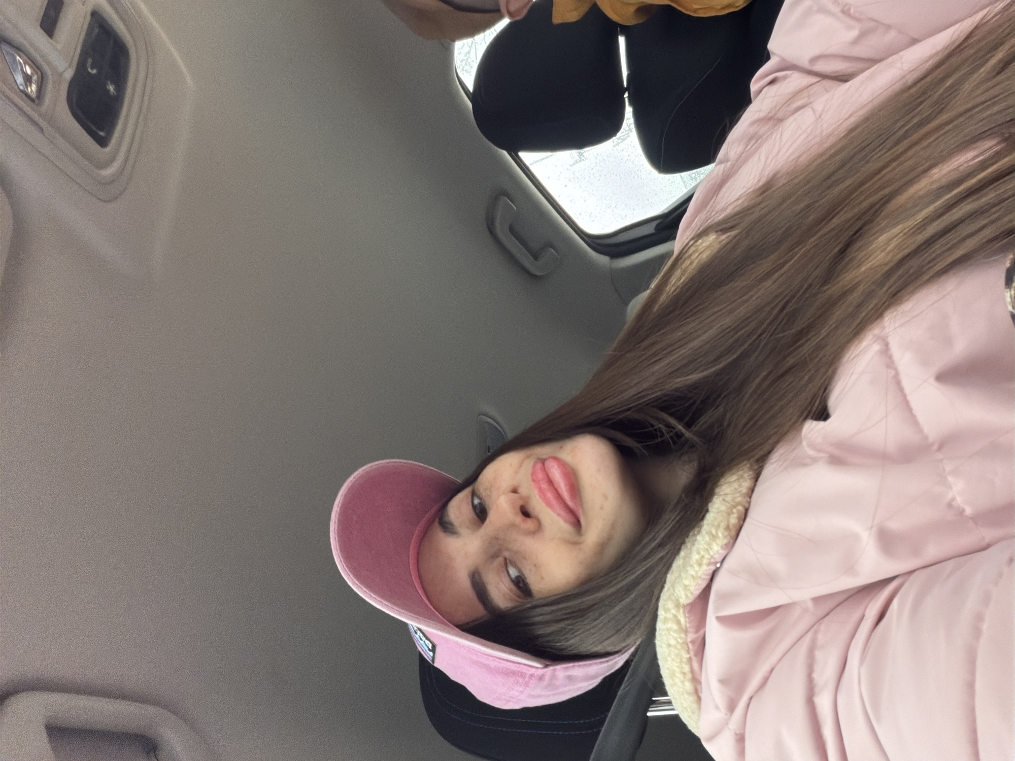 A woman wearing a pink baseball cap and a pink puffer jacket is seated in a vehicle. The interior of the vehicle is visible, including the ceiling, seats, and a sunroof. The environment outside the sunroof appears to be wet, possibly raining. The exact location in Saint Petersburg, Russia, cannot be determined from the image.
