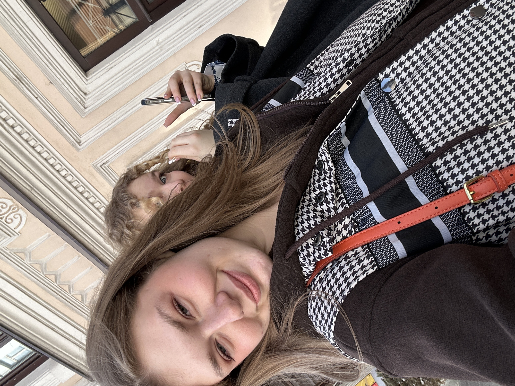 The image depicts two young adult females in an outdoor urban environment in Kazan, Russia. The primary subject, in the foreground on the left, has long, light brown hair and a fair complexion with a distinct reddish hue on her cheeks. She displays a slight smile and makes direct eye contact with the viewer. Her attire includes a dark brown fleece-like hooded garment, layered over a black and white houndstooth pattern shirt or jacket. An orange strap, consistent with a shoulder bag, is visible across her upper torso, featuring a metallic buckle.
The secondary subject, situated behind and to the right of the primary subject, is partially visible. She possesses curly blonde hair and blue eyes. Her right hand firmly grasps a dark-colored smartphone in a vertical orientation; her fingers are manicured with a white tip (French manicure). Her left hand is raised to the vicinity of her left ear. She wears a dark grey overcoat, beneath which a patterned blouse is discernible.
The background consists of a multi-story building facade, light in color, appearing cream or beige. It features distinct white architectural moldings, cornices, and window surrounds. Specific decorative elements include a scrolled ornament on the upper left and a repeating geometric pattern, resembling a chevron or stepped design, within the upper molding. A window with a dark brown frame is also visible. The exact street address or specific building name within Kazan cannot be determined from the photographic evidence.