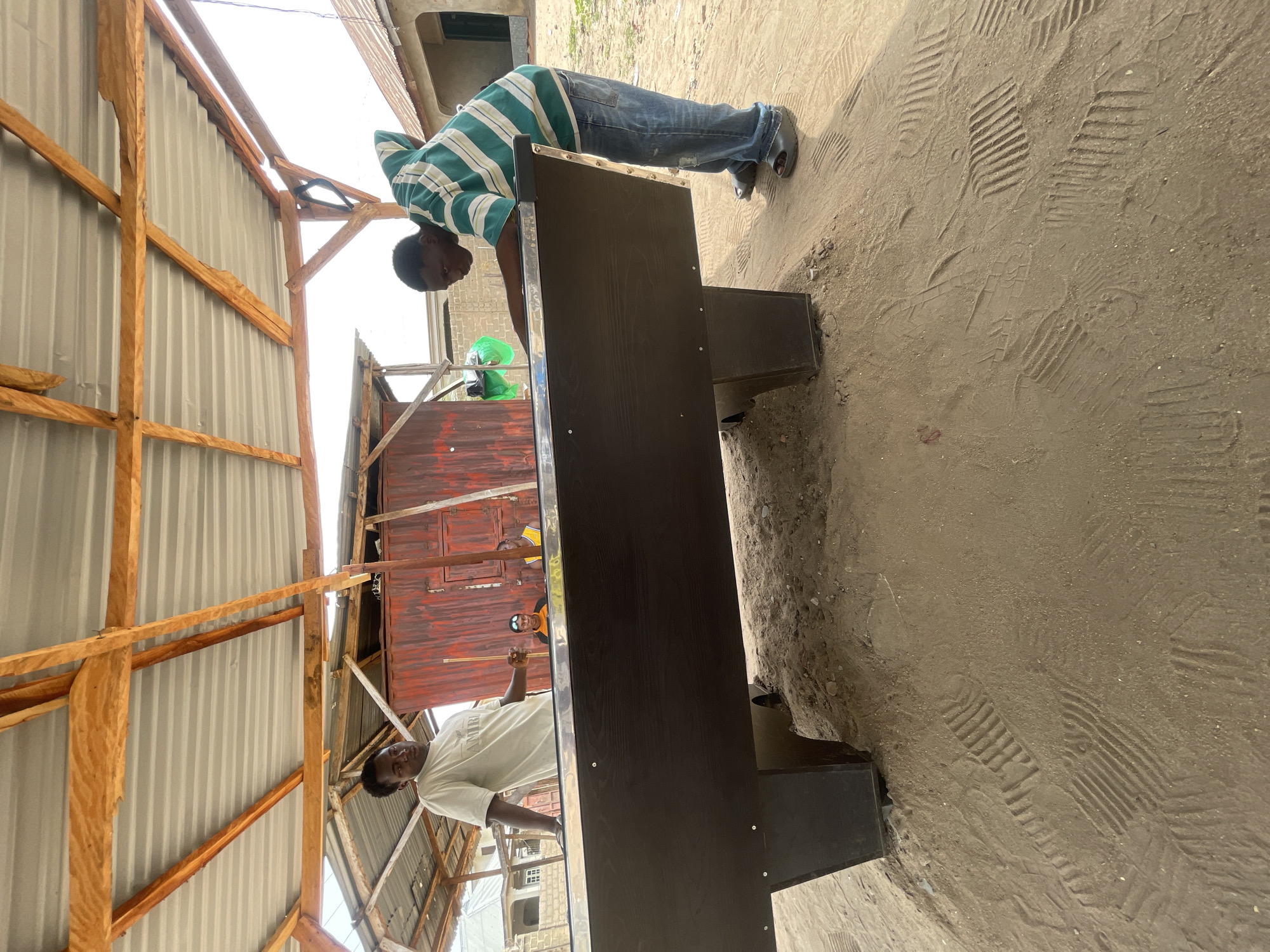 The image shows two men working on a dark-colored rectangular object with metallic trim. One man, wearing a striped shirt and jeans, leans over the object, appearing to adjust or examine it. The other man, wearing a white shirt, stands near the edge of what appears to be a red-painted building or enclosure, holding a tool of some kind.
The location seems to be a partially constructed or open-air structure, possibly in a somewhat undeveloped area. The roofing appears to be corrugated metal, supported by wooden beams. The ground consists of dirt or sand with numerous footprints visible.
Given the prompt constraints, I do not have explicit, forensically-acquired geographic location data for this photograph.