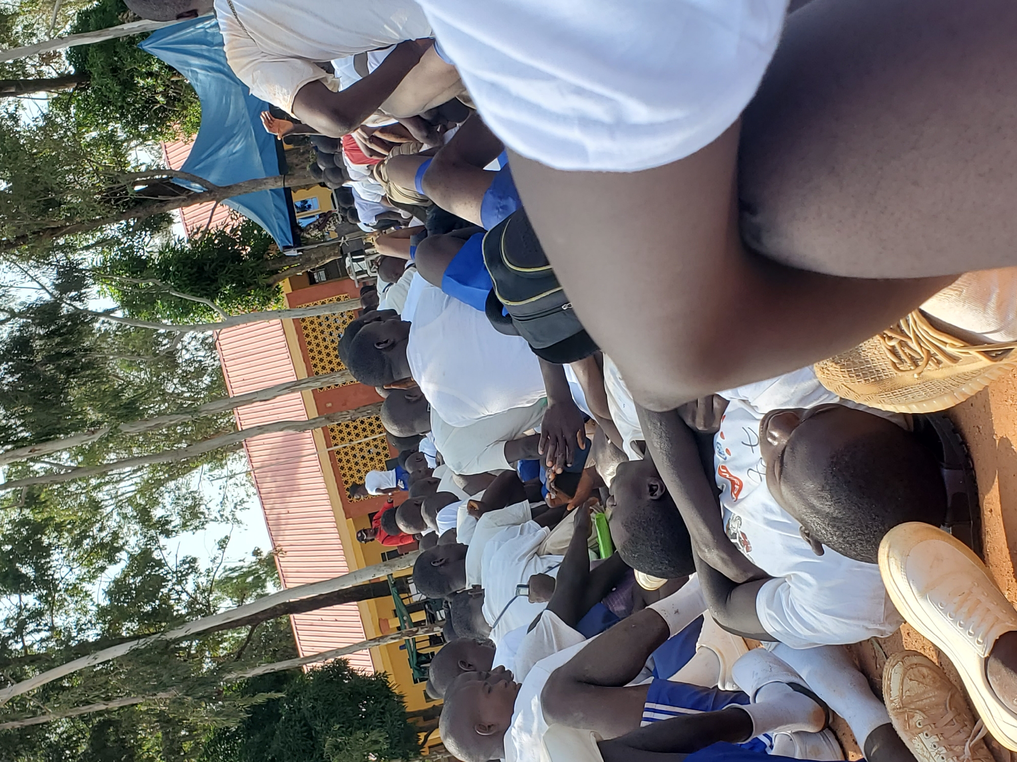 In Barakin Rafin Gora, Nigeria, a group of young men are seated on the ground, appearing to be gathered outdoors near a building with a red corrugated metal roof and yellow and orange patterned walls. Tall, slender trees with green foliage form a backdrop, and a blue tent is visible in the background. Most of the individuals are wearing white t-shirts and shorts, with some wearing white socks and athletic shoes. The perspective is close-up, with some individuals in the foreground and others further back. One person is lying down on their back, looking upwards. Several people are holding or interacting with cell phones. The overall impression is a casual gathering or assembly.