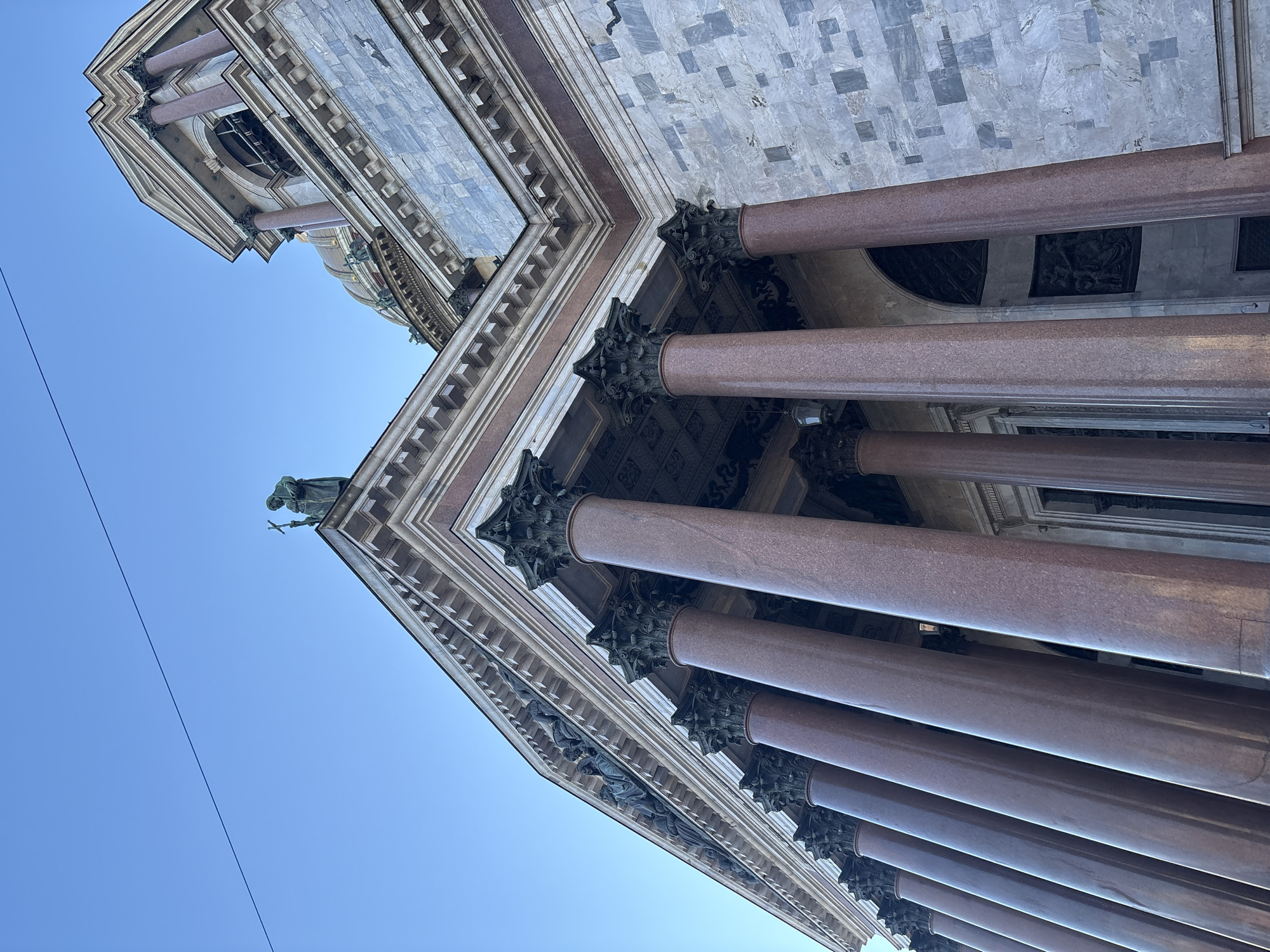 This image depicts a low-angle view of a portion of St. Isaac's Cathedral in Saint Petersburg, Russia, under a clear blue sky. The foreground is dominated by a series of massive, polished reddish-brown granite columns, each topped with a dark, ornate Corinthian capital featuring acanthus leaf designs. Above these columns, a classical pediment is visible, containing a detailed bas-relief sculpture depicting multiple draped human figures.
On the roofline at the corner of the building, a dark-colored statue of a robed figure holding a cross is prominently positioned against the sky. To the right of the main columned portico, a section of the cathedral's wall is constructed from light-colored, irregularly patterned stone blocks, possibly granite or marble. Further to the right and partially obscured, a decorative upper section features smaller columns, an arched window or opening, and architectural detailing.
In the background, a segment of the cathedral's large, golden dome is visible, with sculpted figures appearing at its base. A thin, dark horizontal wire stretches across the upper left portion of the clear blue sky. No human figures or discernible actions are present within the frame.