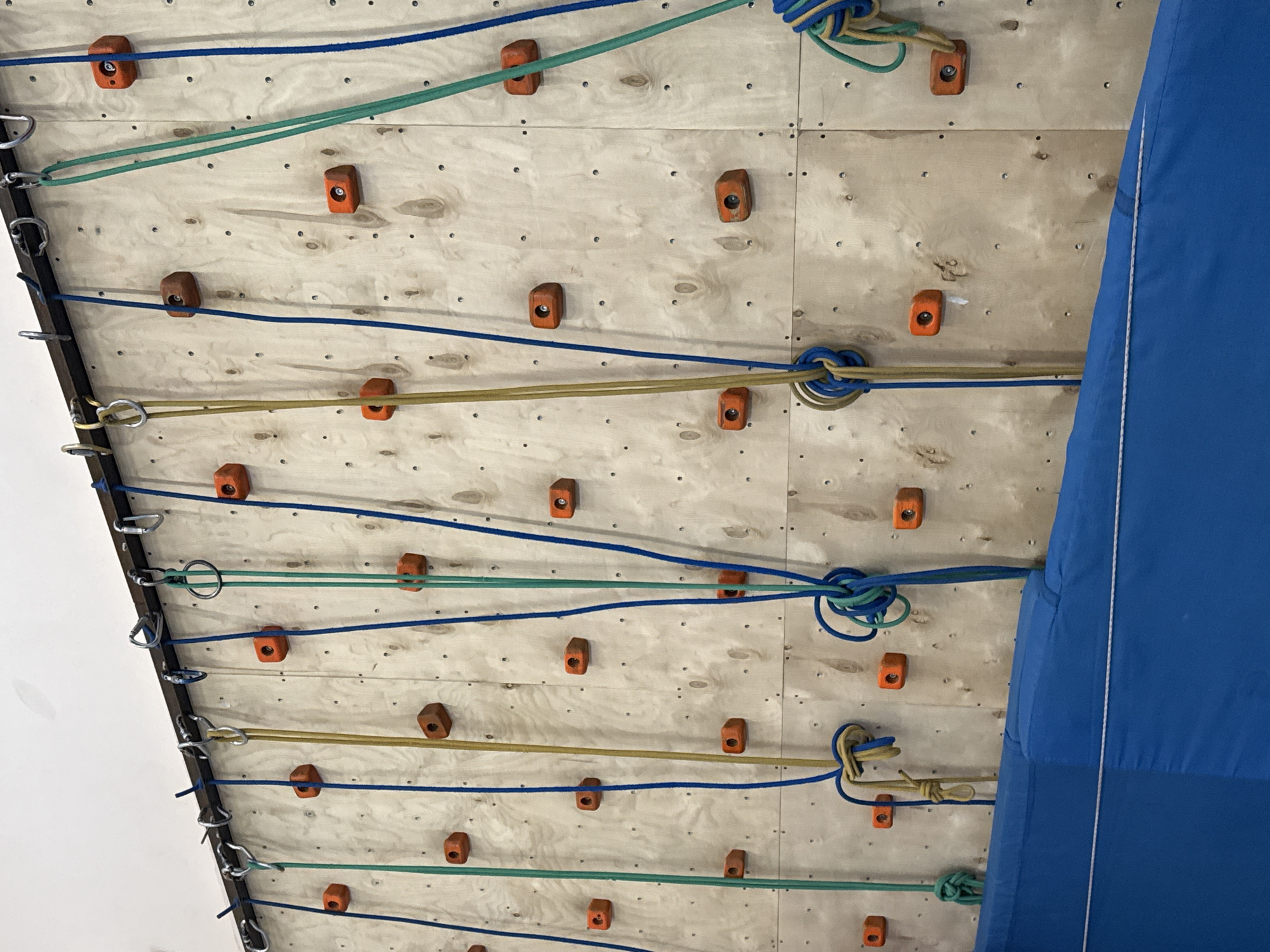 The image depicts a climbing wall, constructed of wooden panels with bolted-on orange climbing holds. Several ropes in blue, green, and yellow are strung across the wall, passing through metal carabiners or rings attached to the left edge. The right side of the wall is partially obscured by a blue padded mat. The wall appears to be indoor, as evidenced by the adjacent white wall. There are no people visible. The location is unknown, and there is no information within the image to suggest it is in Usolye, Russia.