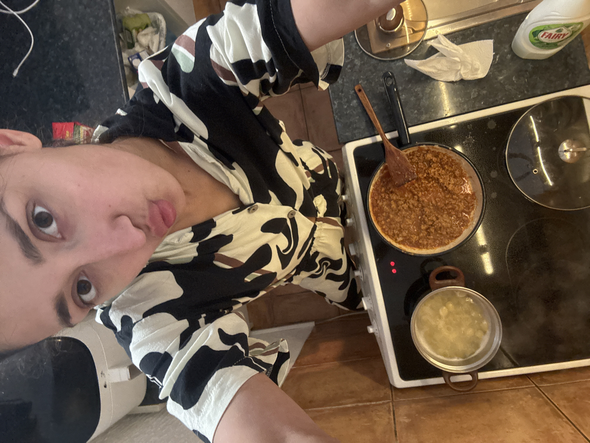 The image captures a woman taking a selfie in what appears to be a kitchen. She is wearing a patterned black and white dress. In the background is an electric stove top with two pans on it. One pan contains a brown sauce, possibly meat-based, and a wooden spoon rests in it. The other pan appears to contain a light-colored liquid with some solid pieces. To the upper right is a bottle of Fairy dish soap and a crumpled white paper towel. The kitchen features dark countertops, and the floor is tiled. The location appears to be a domestic setting. It is not possible to determine if this picture was taken in Sutomore, Montenegro from the given information.
