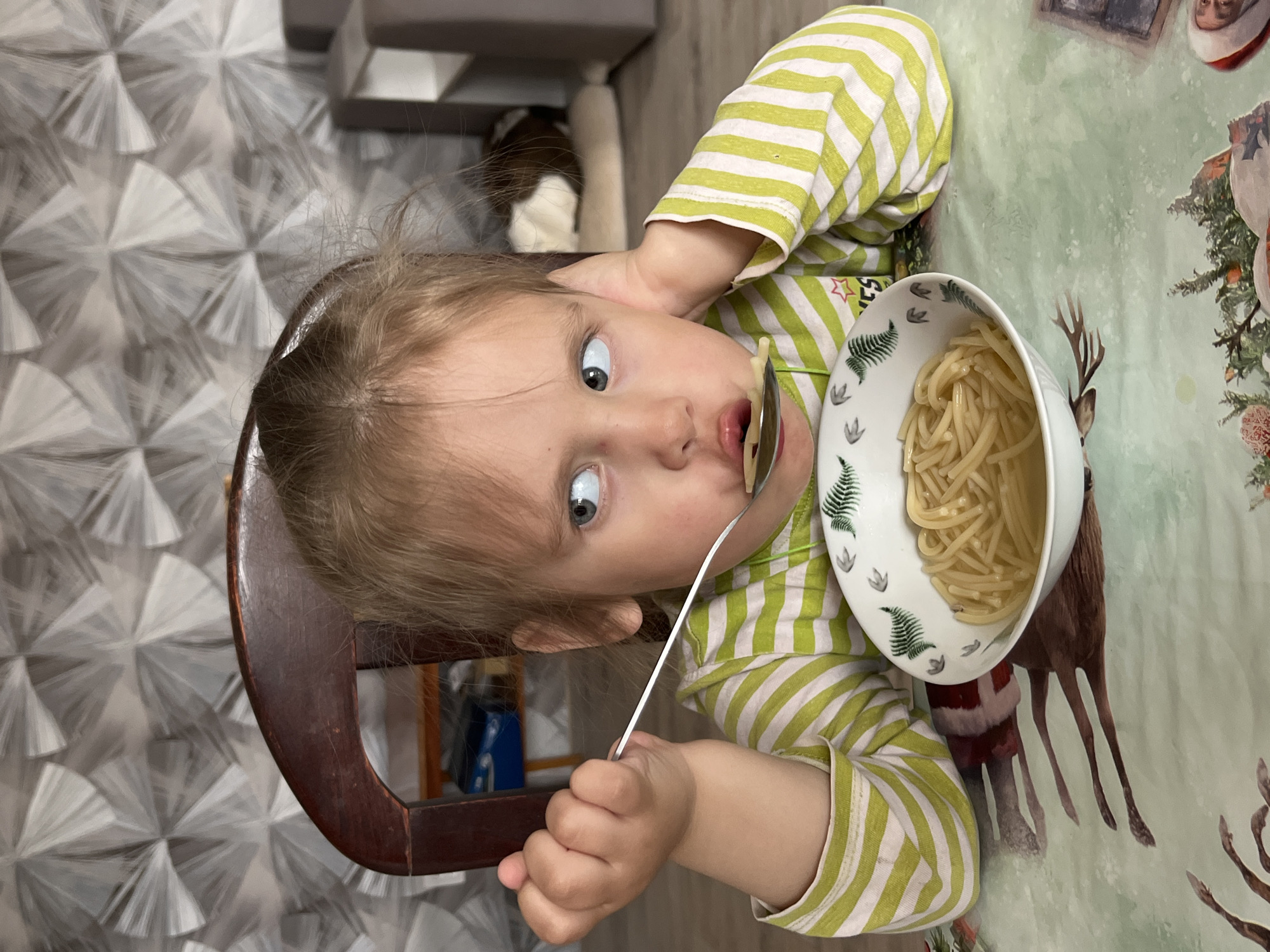 A young child with light hair and blue eyes is captured in a close-up, eating pasta at a table indoors. The child wears a light green and white striped long-sleeved shirt and has a wide-eyed, perhaps surprised or focused, expression as a spoonful of light-colored pasta is held to their mouth. The pasta is in a white bowl adorned with green fern and paw print designs. The child is seated in a dark wooden chair.
The table is covered with a festive tablecloth featuring a light green background with holiday-themed illustrations of reindeer and Christmas trees, hinting at a winter or holiday season. In the background, behind the child, there is a distinct wall with a silver-white geometric pattern resembling radiating fans. Further back and out of focus, a brown and white cat is partially visible, along with some domestic furniture or shelving. The scene depicts a mealtime moment in a home, located in Chaikovsky, Russia. There is no visible text.