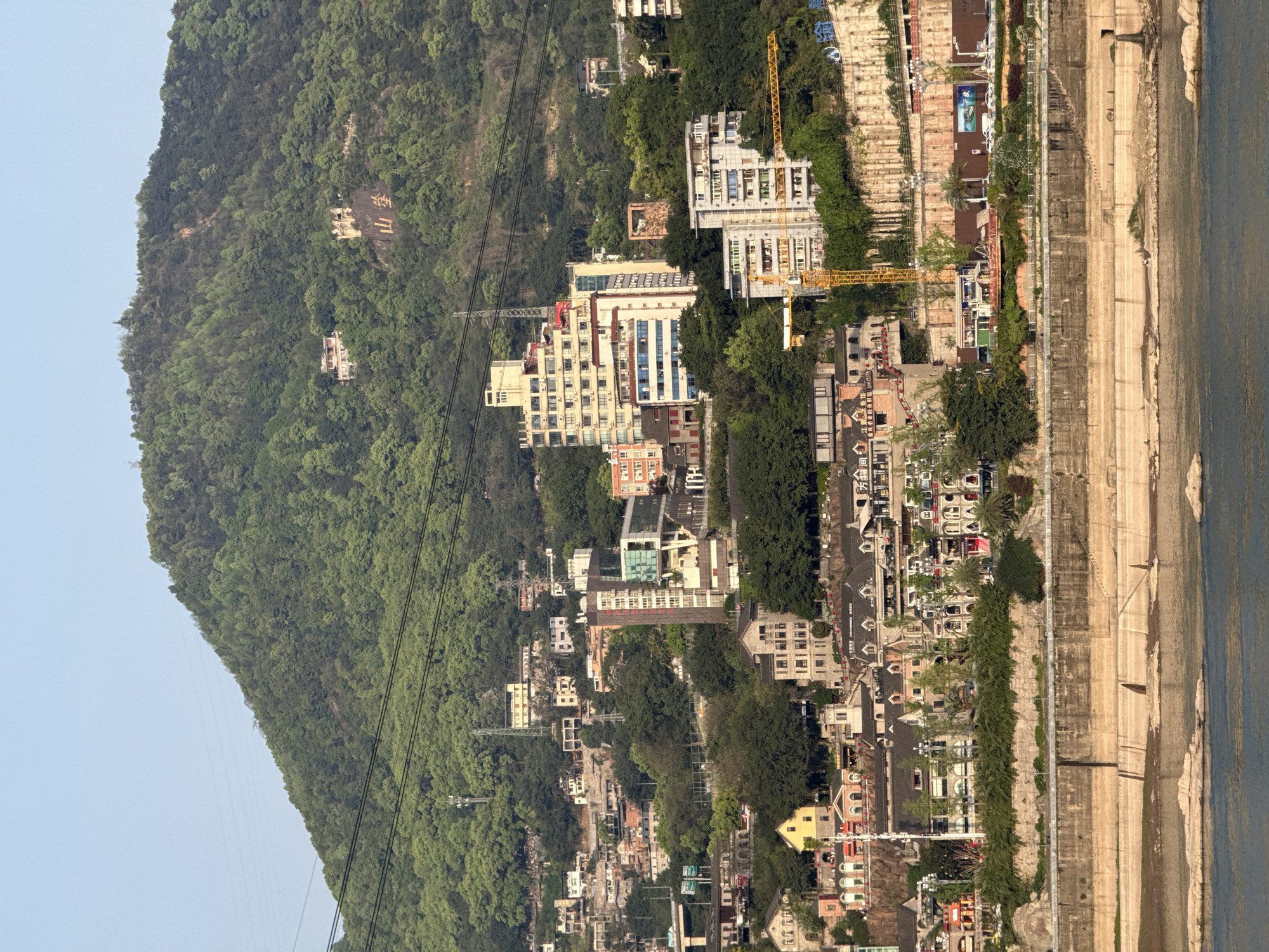 The image captures a landscape along a river in Yuzhong District, China. A densely forested mountain dominates the left side, with some construction and development. Buildings of varying heights and architectural styles line the riverbank. Construction cranes are visible near newer buildings. Below the buildings is an embankment of several levels and then the river. Power lines run across the mountain side.