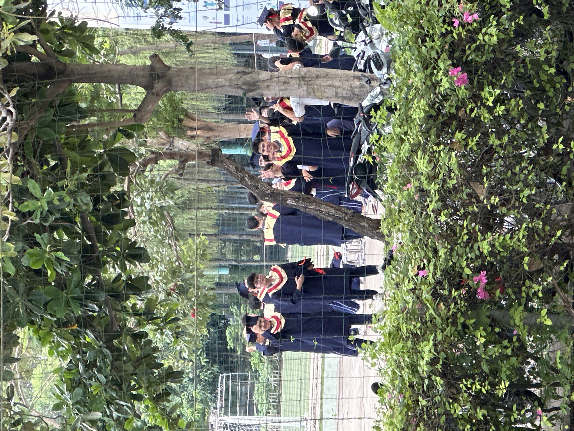 The image shows a group of graduates in Đồng Nai, Vietnam, seen through a chain-link fence and trees. The graduates are wearing graduation gowns and stoles, and some are waving. Parked motorcycles are visible in the background. The environment includes trees and flowering bushes.
